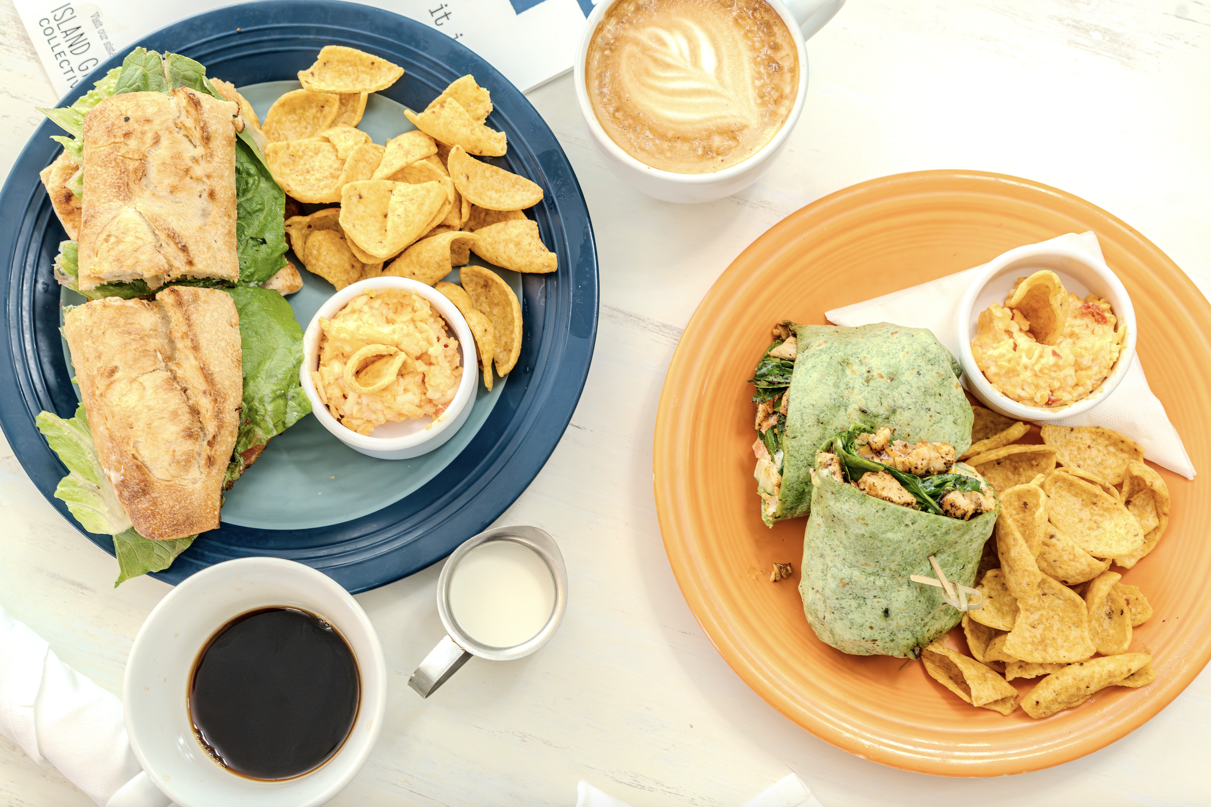 Plates of sandwiches with chips and sides, a cup of coffee, and a latte.
