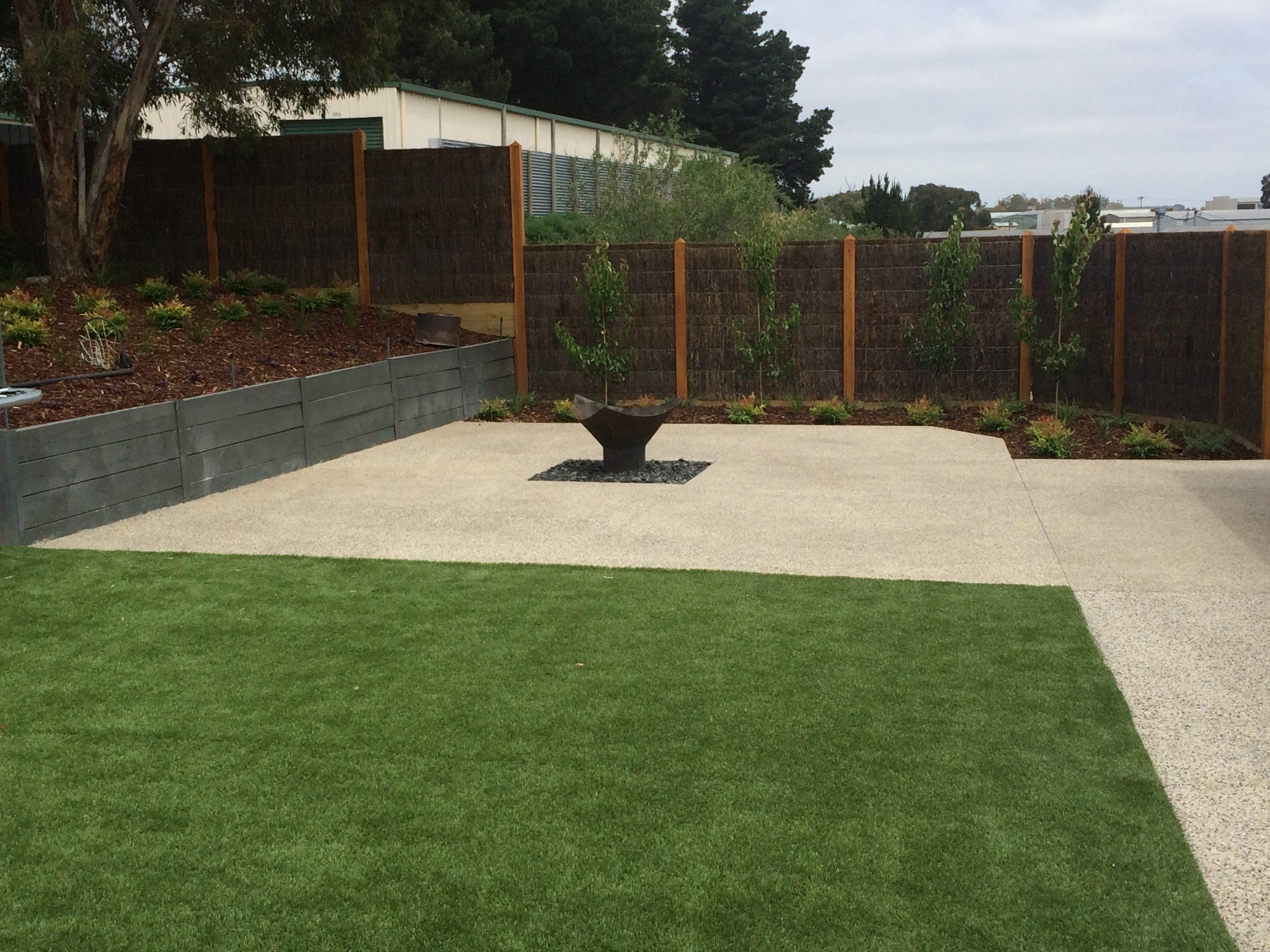 Synthetic turf, retaining wall with garden bed, exposed aggregate, fire pit and brush panel fencing with exposed posts