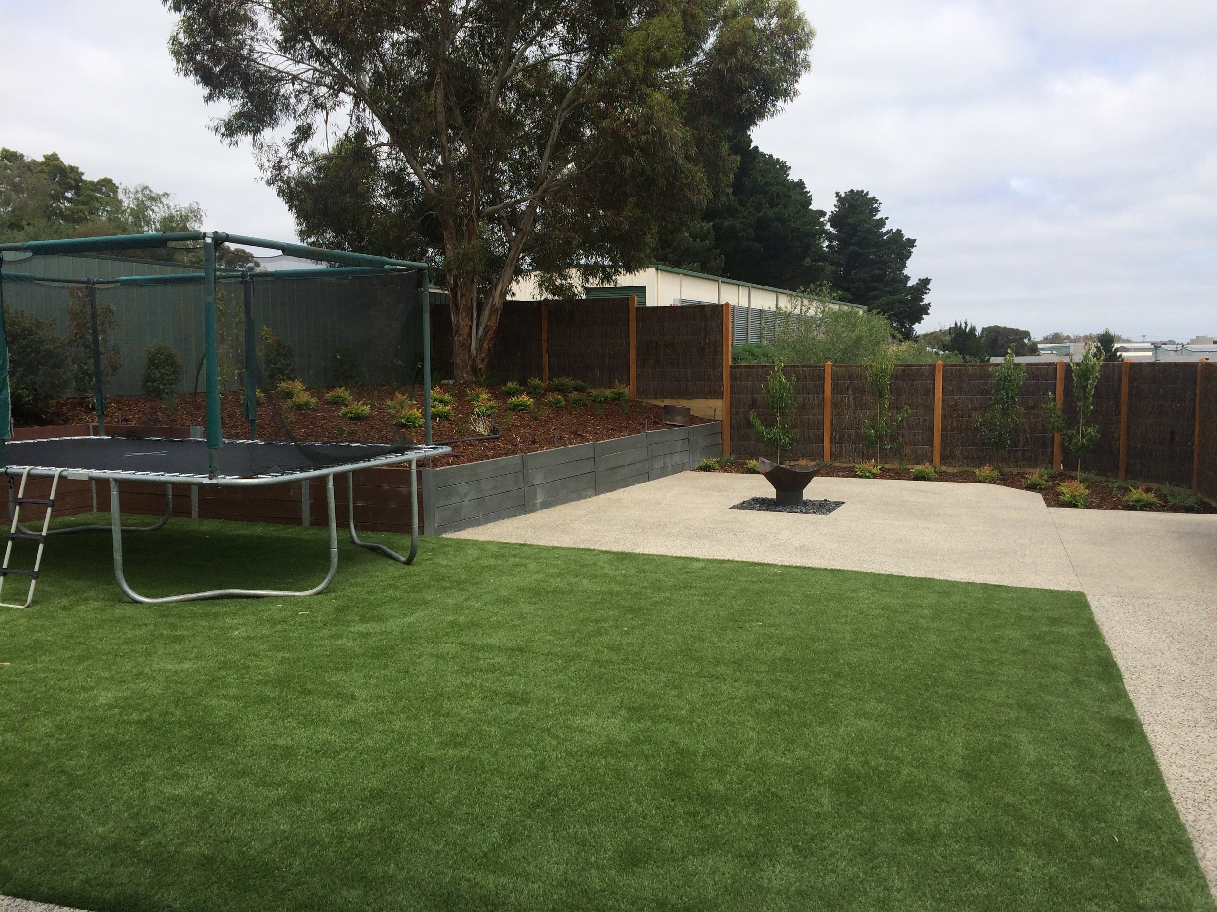 Synthetic turf, retaining wall with garden bed, exposed aggregate, fire pit and brush panel fencing with exposed posts