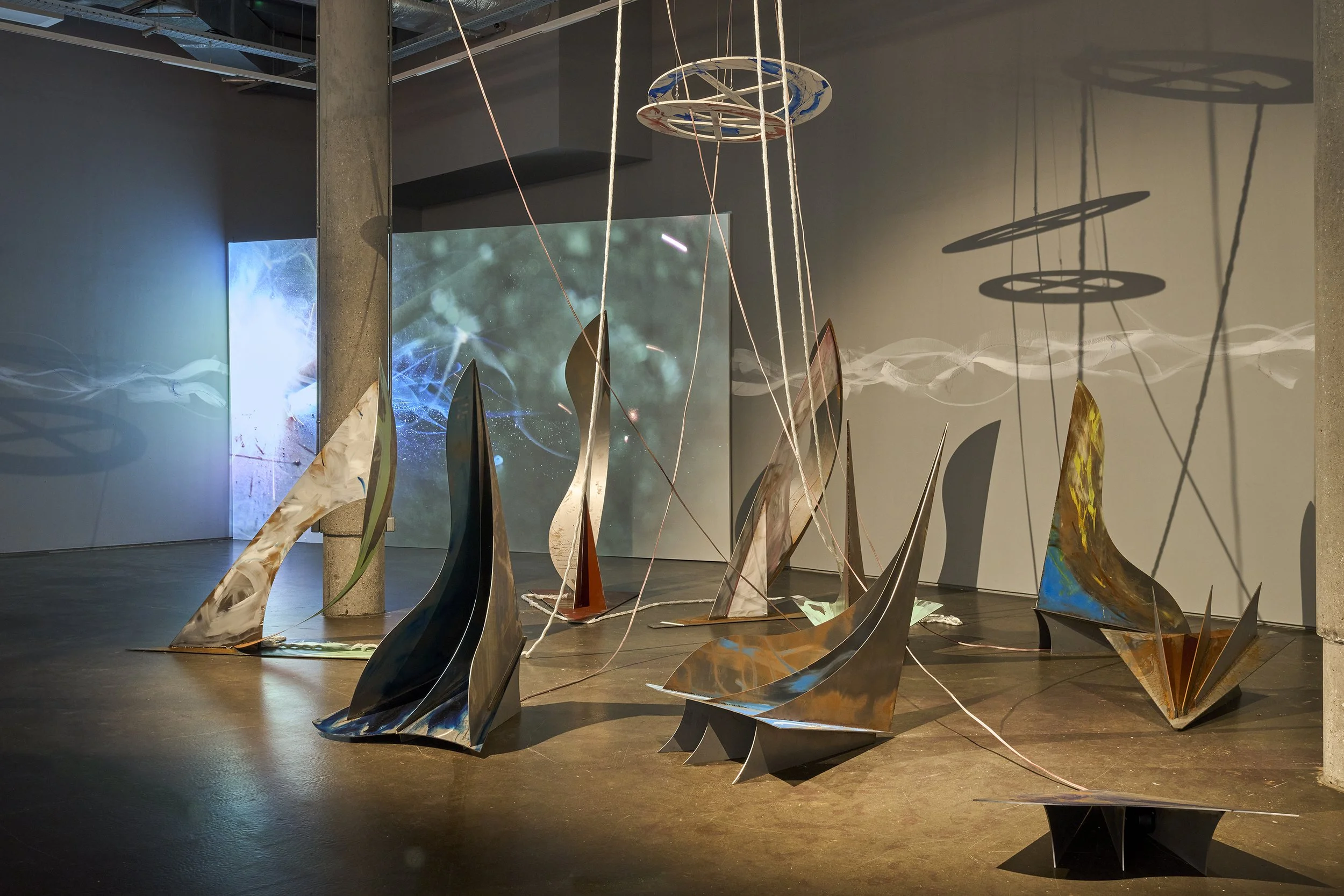 A multi-part sculptural installation of steel sculpture, video and sound by artist Ro Robertson. 9 steel painted sculptures are situated in front of a large screen which shows a close up of welding echoed by the white painted line that spans the wall