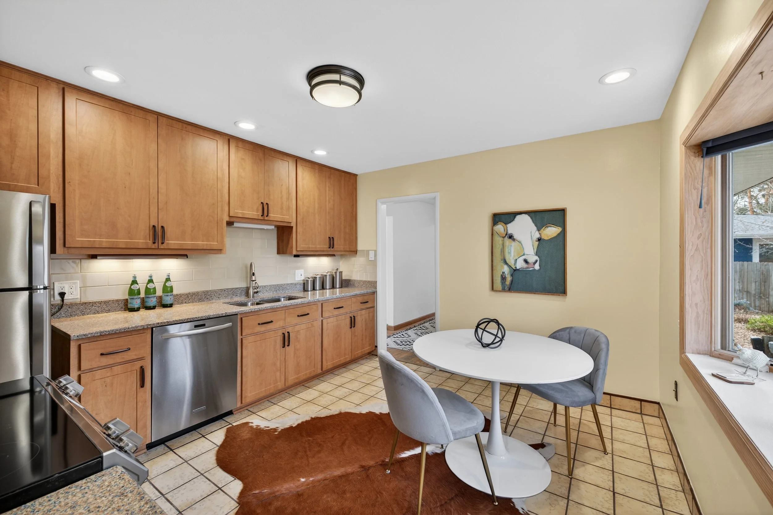 Kitchen with wooden cabinets, stainless steel appliances, a small round dining table with three gray chairs, a cow painting on a yellow wall, window with a view of backyard, tiled floor, cowhide rug.
