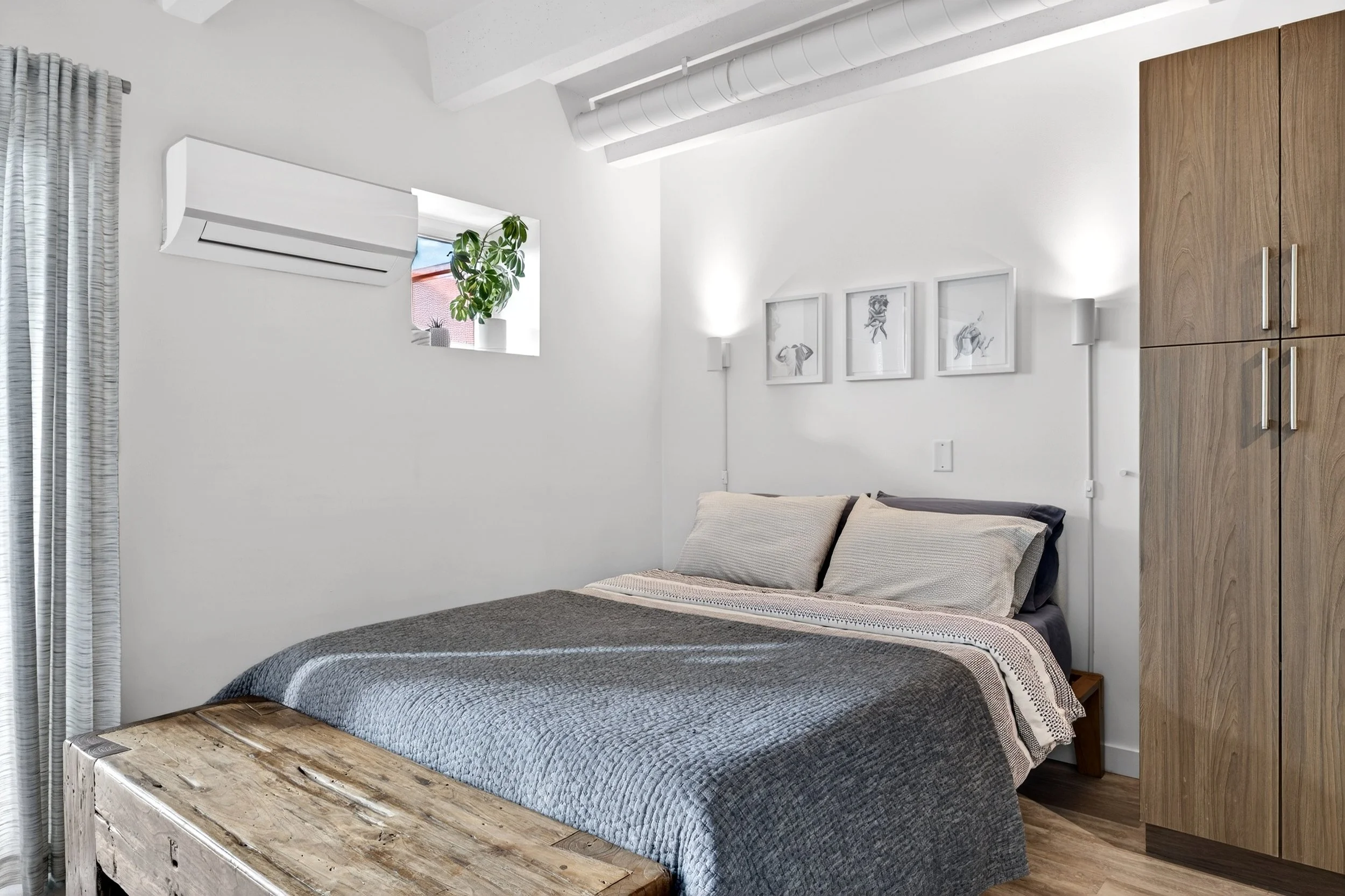 Bright minimalist bedroom with white walls, a wooden wardrobe, a bed with beige and gray bedding, art prints on the wall, and a small window with a potted plant.