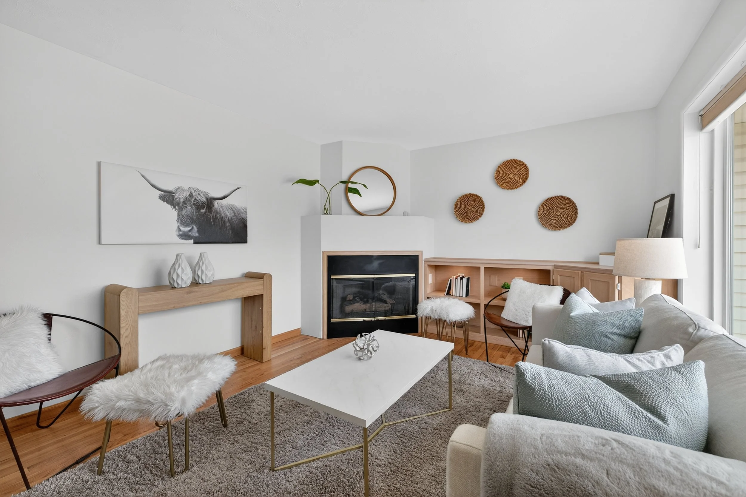 Modern living room with white walls, wooden accents, and a fireplace. Contains a white sofa with pastel pillows, a white coffee table, a cozy chair with faux fur, wall art, and woven baskets on the wall.