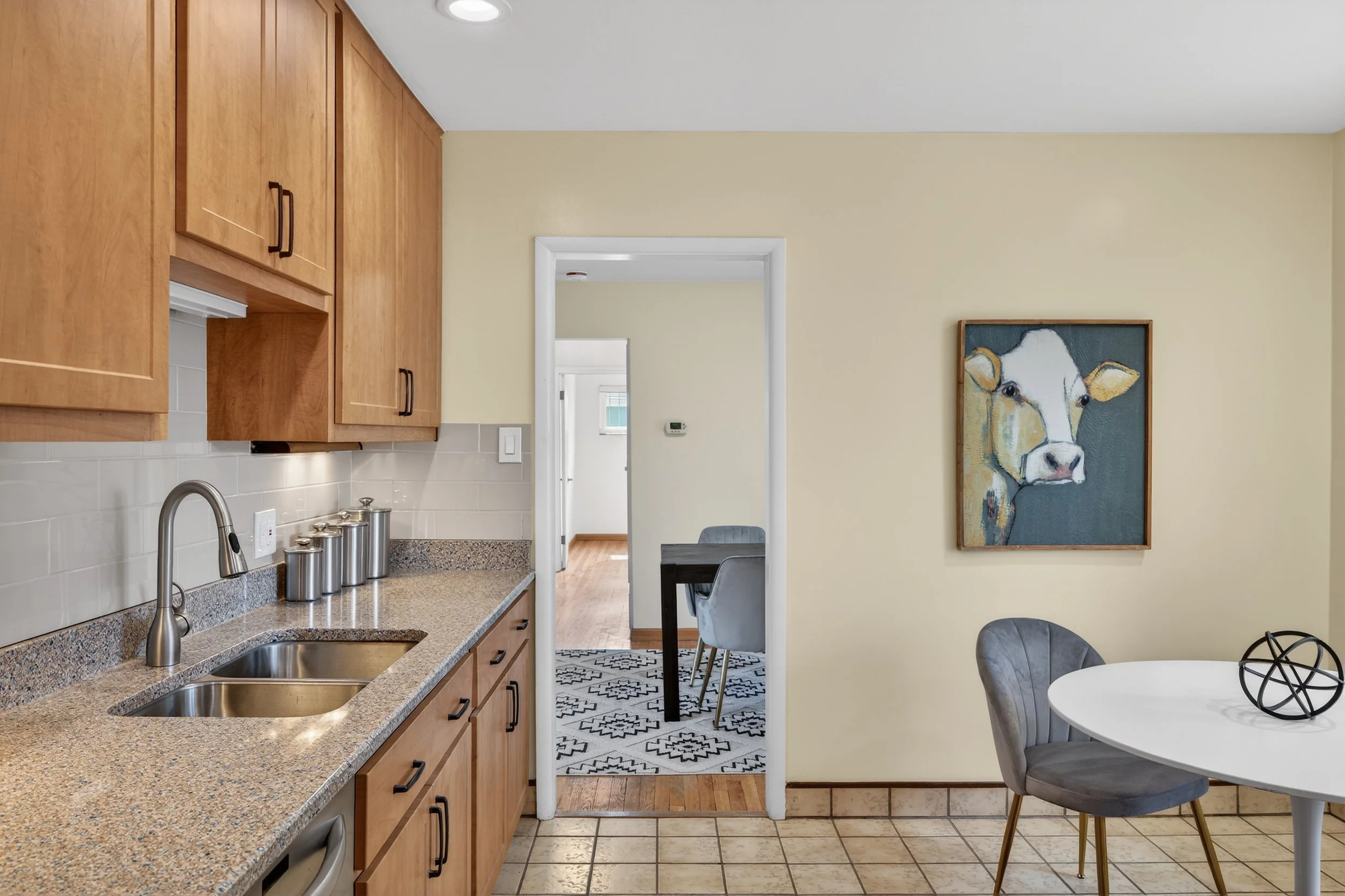 Kitchen with wood cabinets, granite countertop, stainless steel sink, light beige tile backsplash, and a view into dining and living areas with artwork of a cow on the wall.