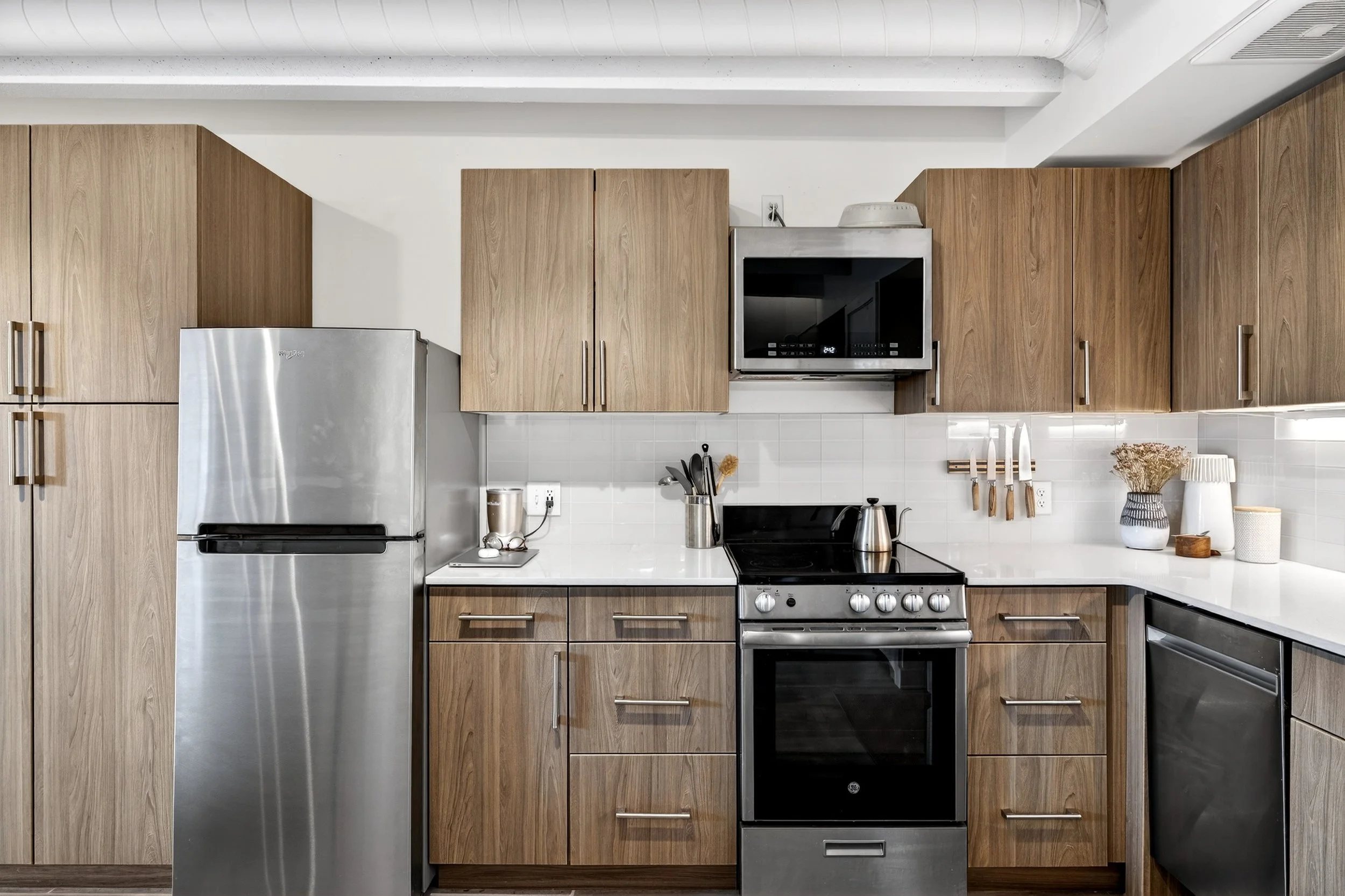 Modern kitchen with wooden cabinets, stainless steel refrigerator, stove, microwave, and decorative items on the white countertop.