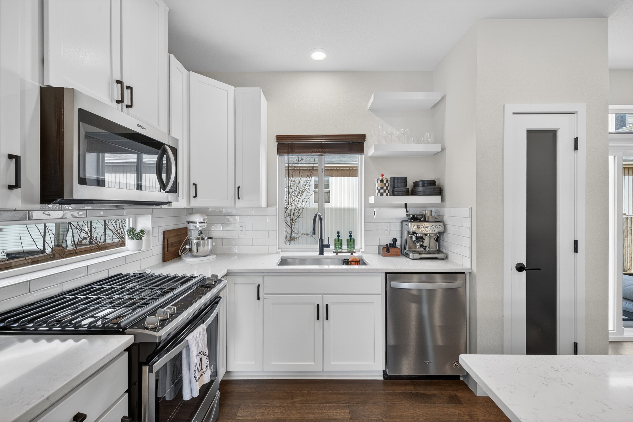Modern kitchen with white cabinets, stainless steel appliances, a window with a brown wooden blind, a black faucet at the sink, open shelves with glassware and plates, and a small countertop espresso machine.