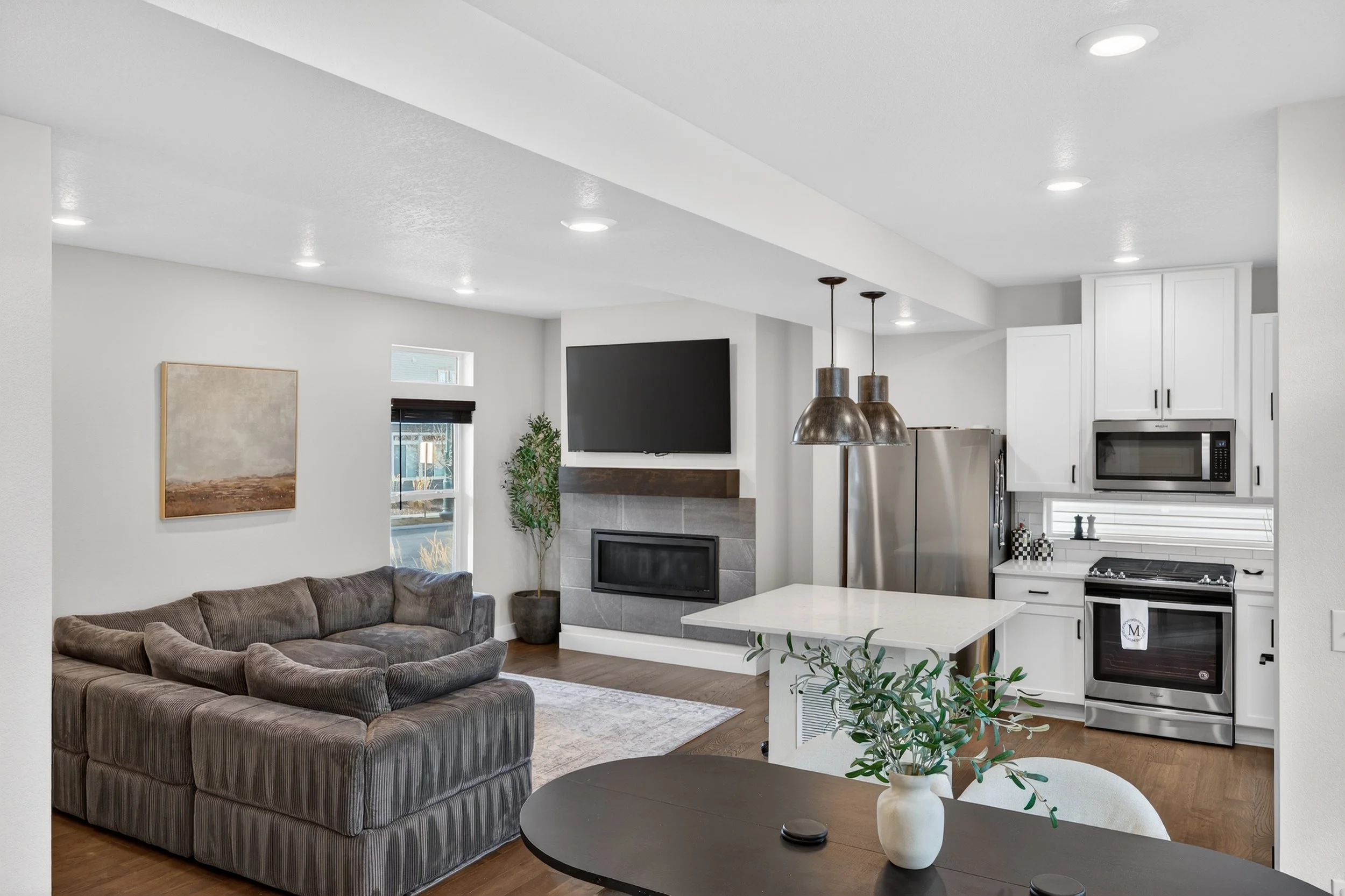 Open-concept living room and kitchen with gray couch, wall-mounted TV, fireplace, potted plant, kitchen cabinetry, stainless steel refrigerator, microwave, oven, and hanging pendant lights.