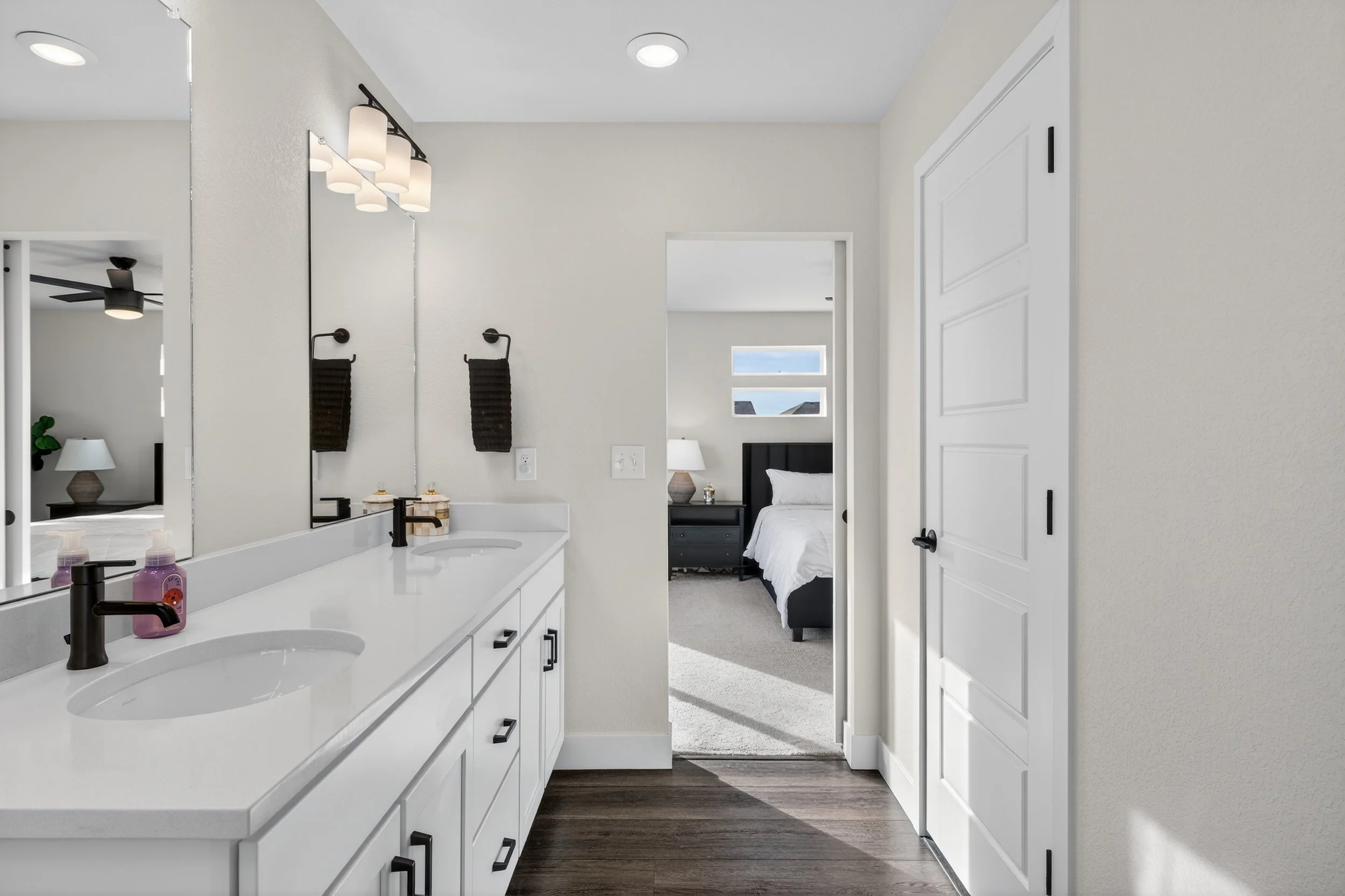 View of a bathroom vanity area with two sinks, a large mirror, black fixtures, black towels, and a bedroom visible through an open doorway in the background, featuring a bed with a dark headboard, white bedding, and a nightstand with a lamp.