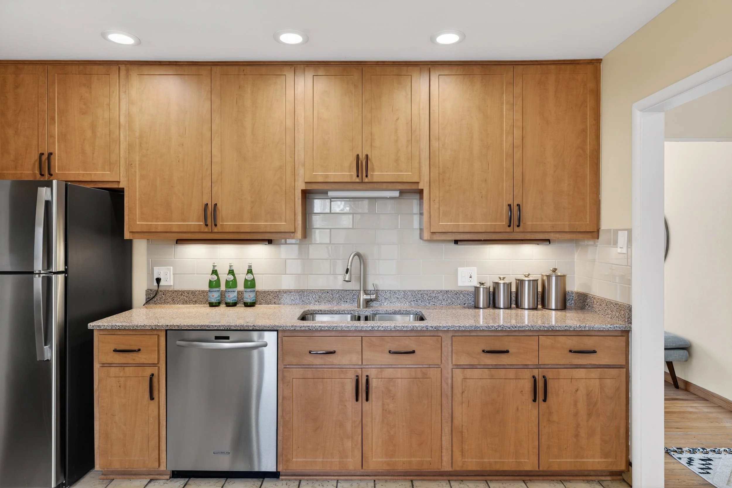 Kitchen with wooden cabinets, stainless steel refrigerator and dishwasher, granite countertop, and beige tile backsplash; three bottles of sparkling water and stainless steel canisters on the counter.