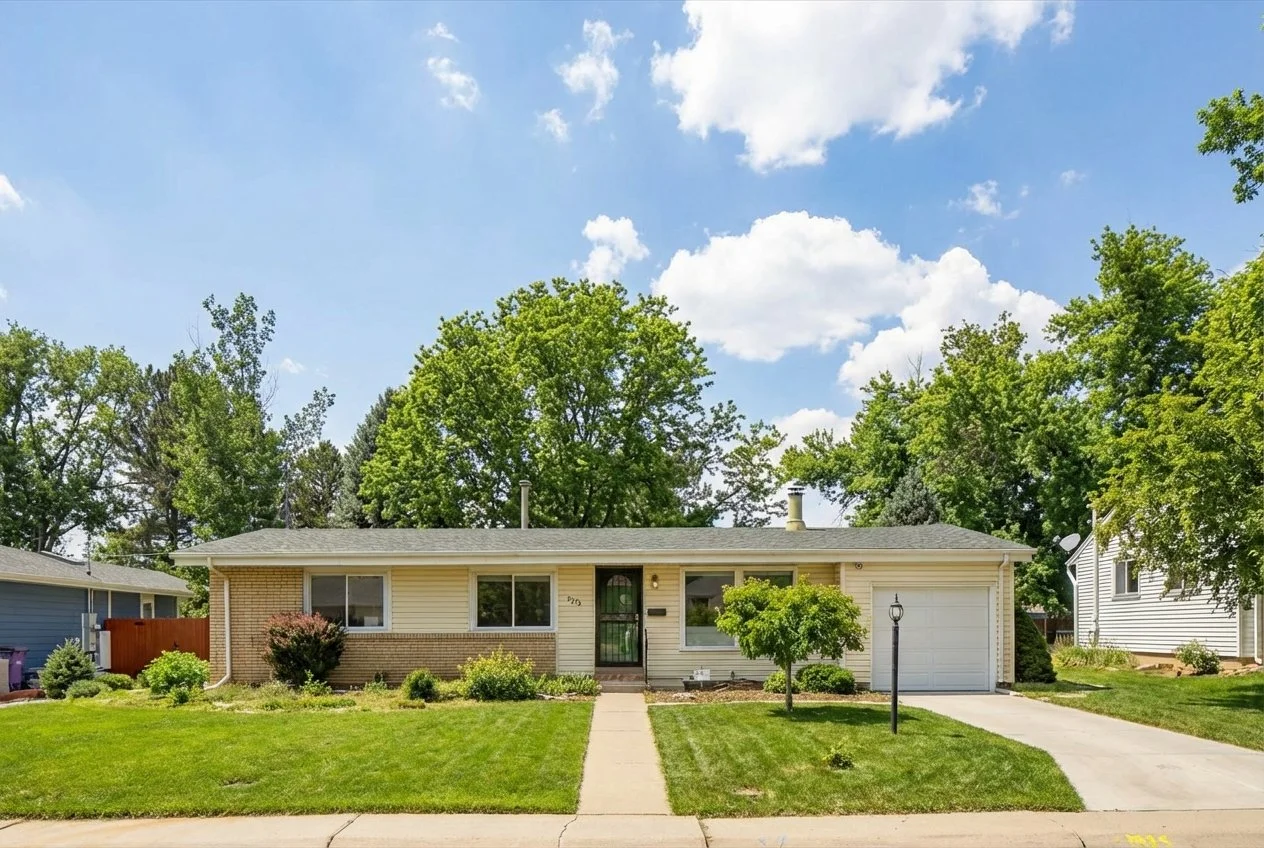 Front view of a single-story house with beige and brick exterior, surrounded by a well-maintained lawn and trees, under a partly cloudy sky.