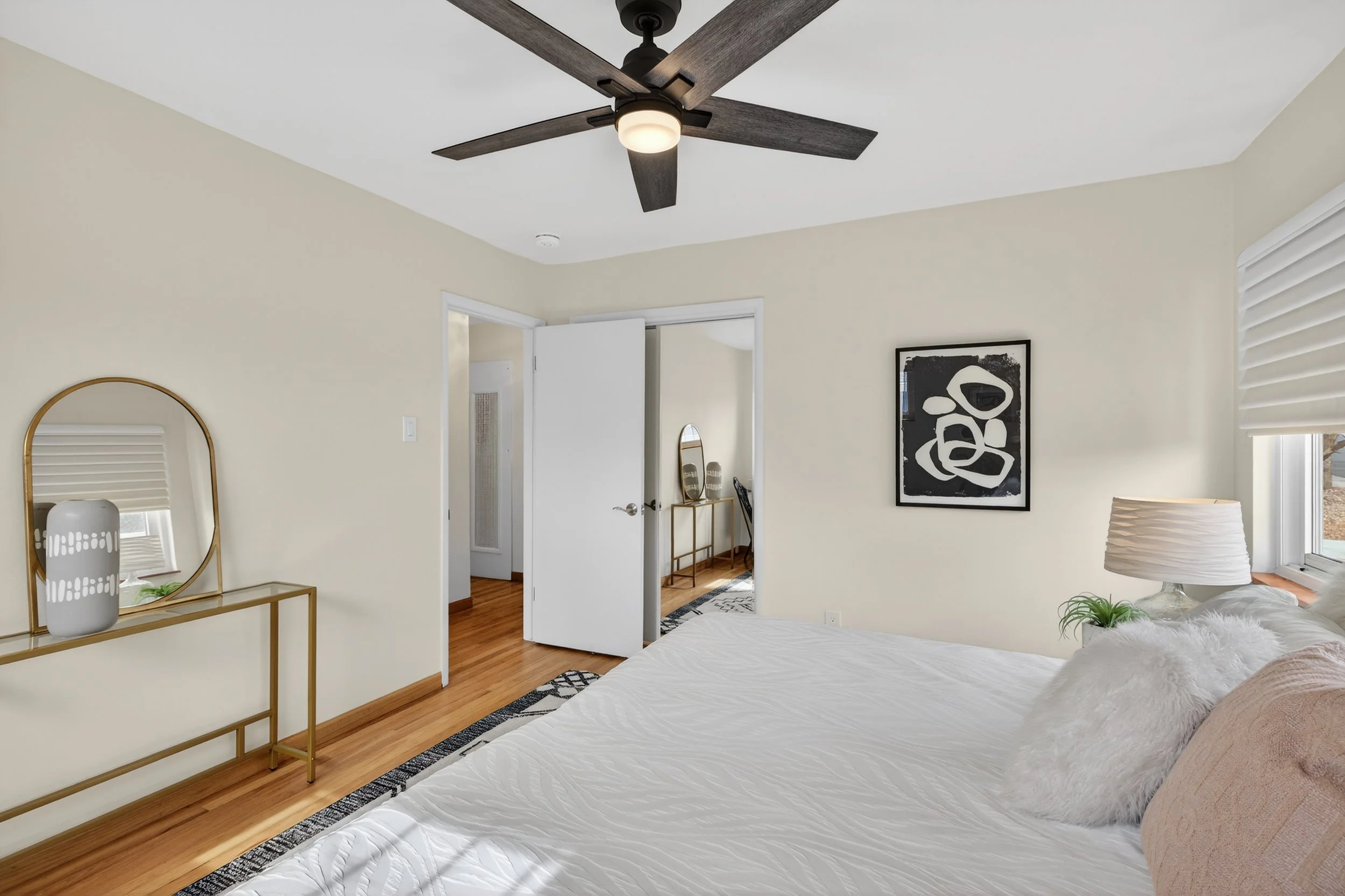 A bedroom with a bed, nightstand, and decorative items. The room has a ceiling fan, a window with a shade, and a door leading to another room. The walls are light-colored, and the flooring is hardwood.