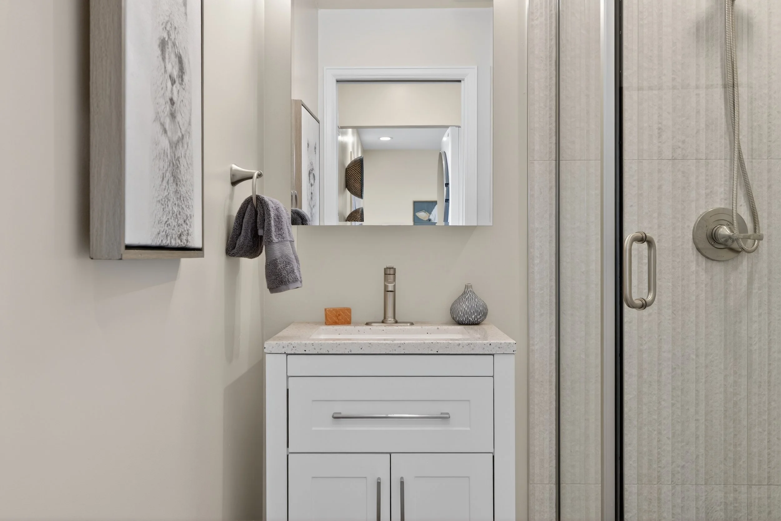 Bathroom with white vanity, speckled countertop, gray towel on wall hook, large mirror, shower with glass door, beige tile walls, and decorative vases on vanity