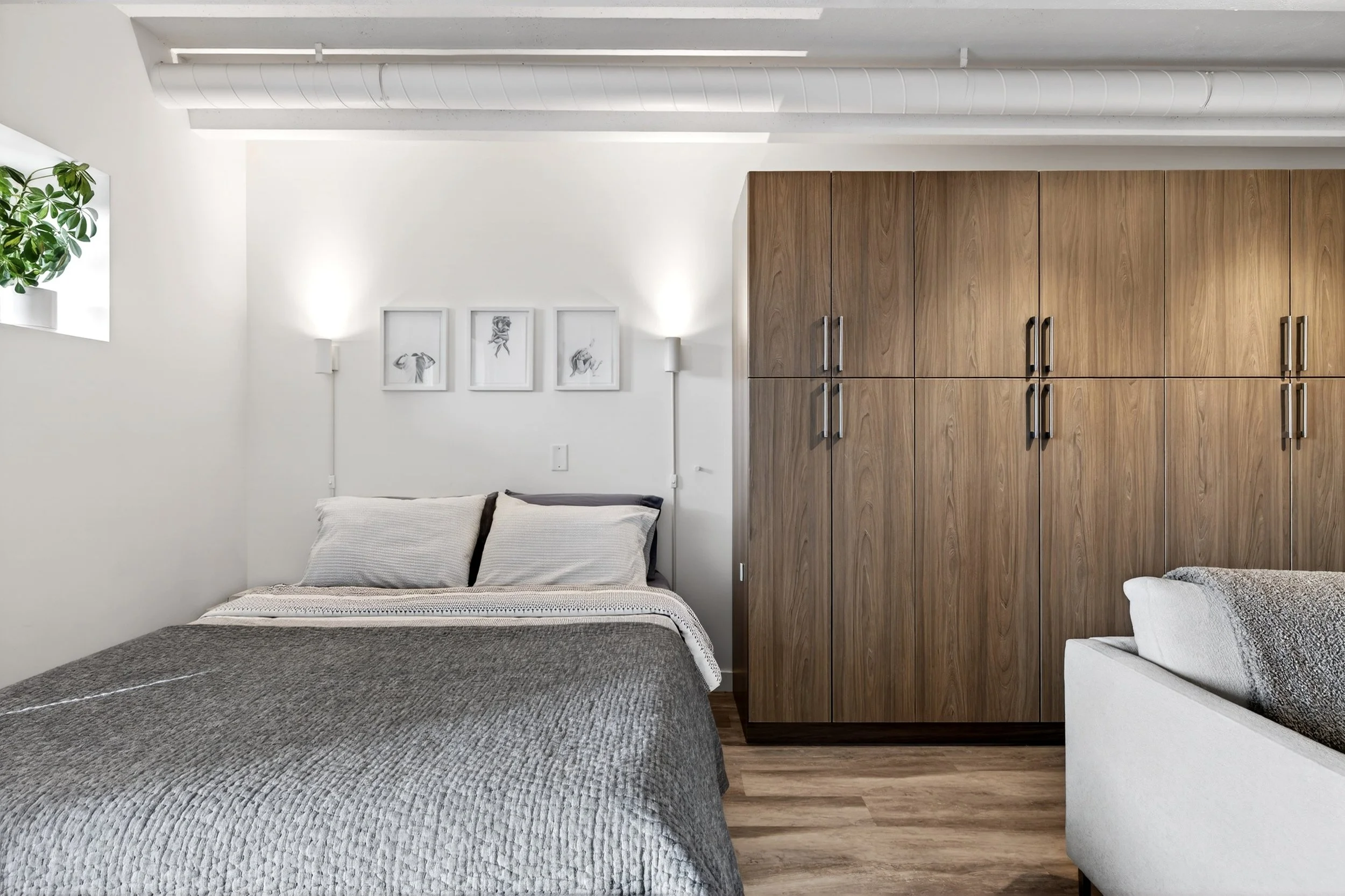 Bedroom with white walls, wooden wardrobe, bed with gray cover, two pillows, wall art, small window with a plant, and hardwood flooring.