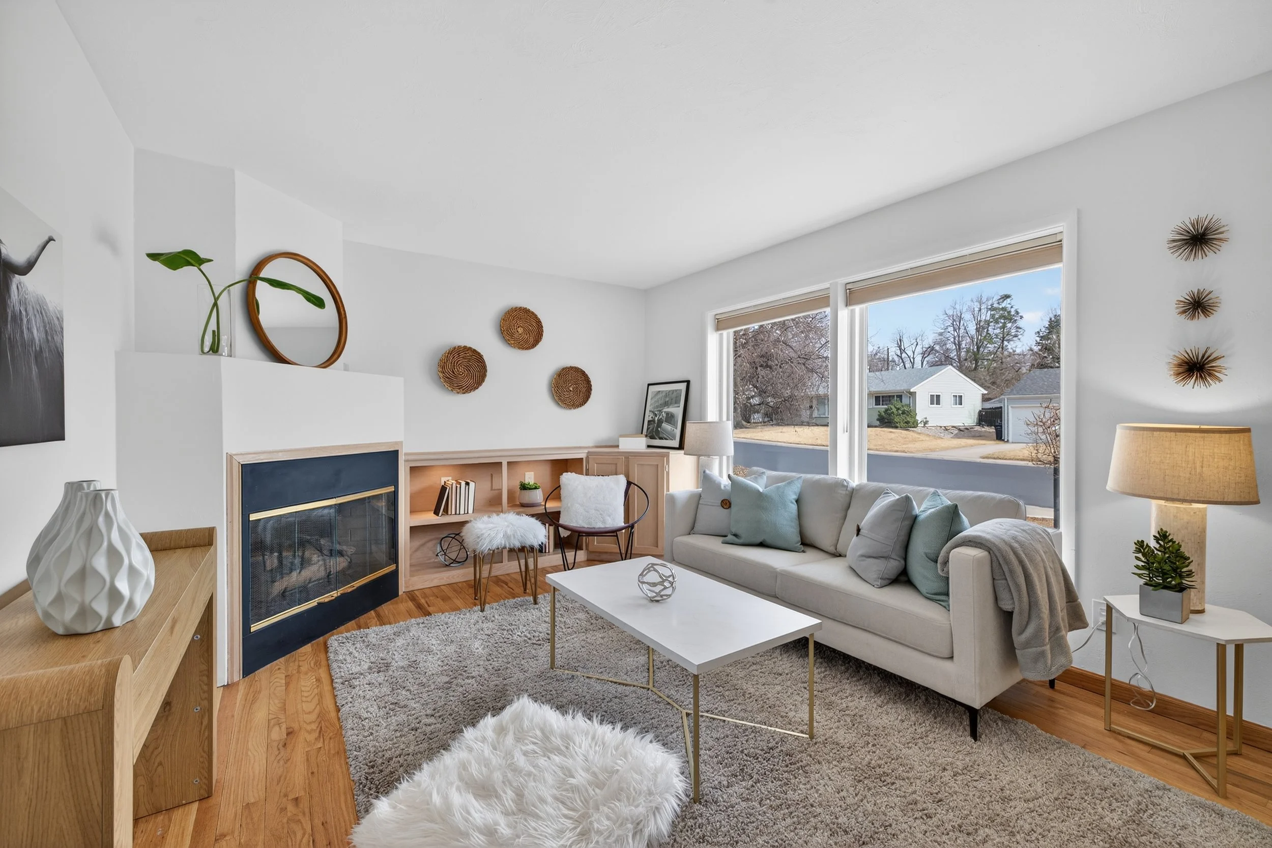 Bright living room with white walls, large windows, a white sofa with pastel pillows, a white coffee table, a cozy furry footstool, a fireplace, and various decorative items including wall art, vases, and plants.