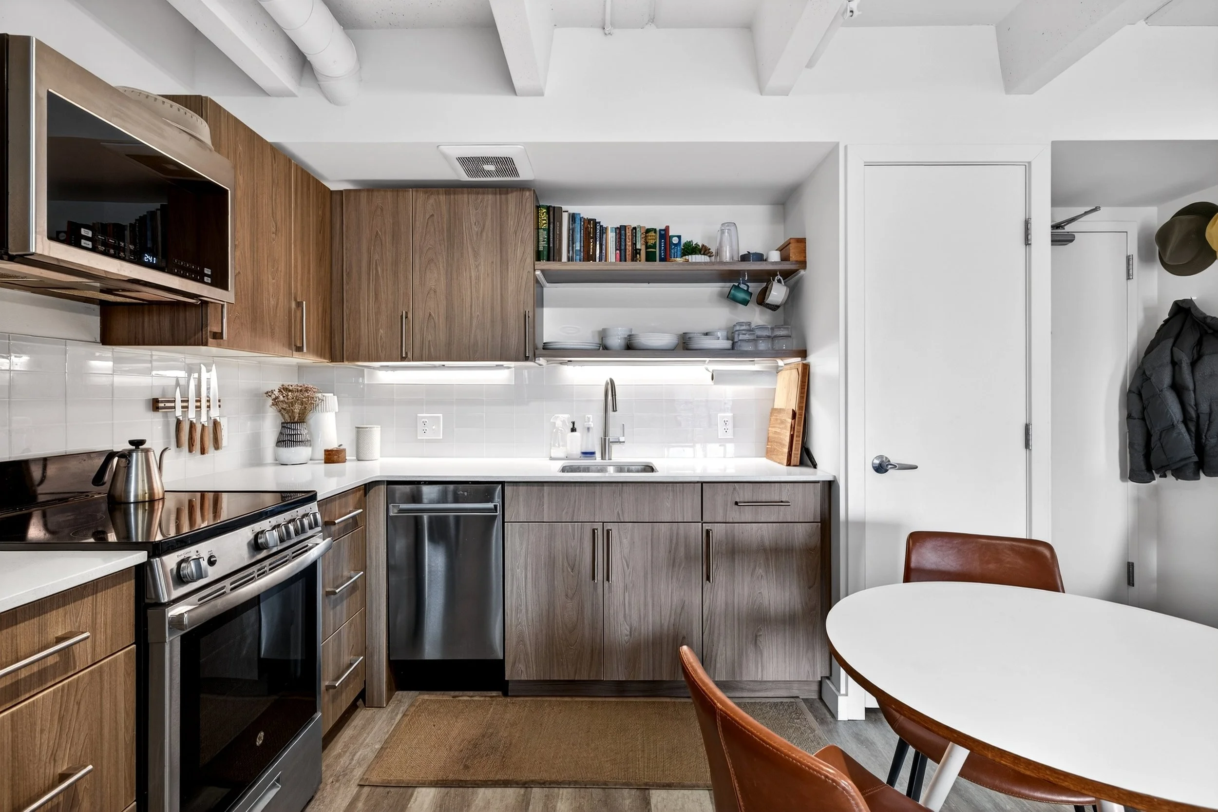 Modern kitchen with wooden cabinets, stainless steel appliances, open shelving with dishes and books, white countertops, small dining table with brown chairs, and a white door on the right.