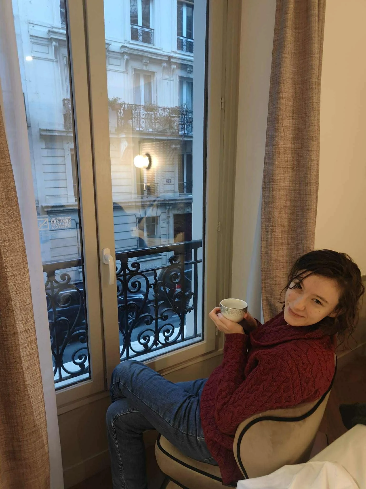 White woman with shoulder-length dark hair, wearing a red jumper and blue jeans, seated indoors near a tall window, holding a cup, with a view of a city street outside.