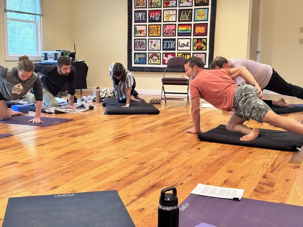 People participating in a yoga or fitness class in a spacious, well-lit room with wooden floors, windows, and colorful artwork on the wall.