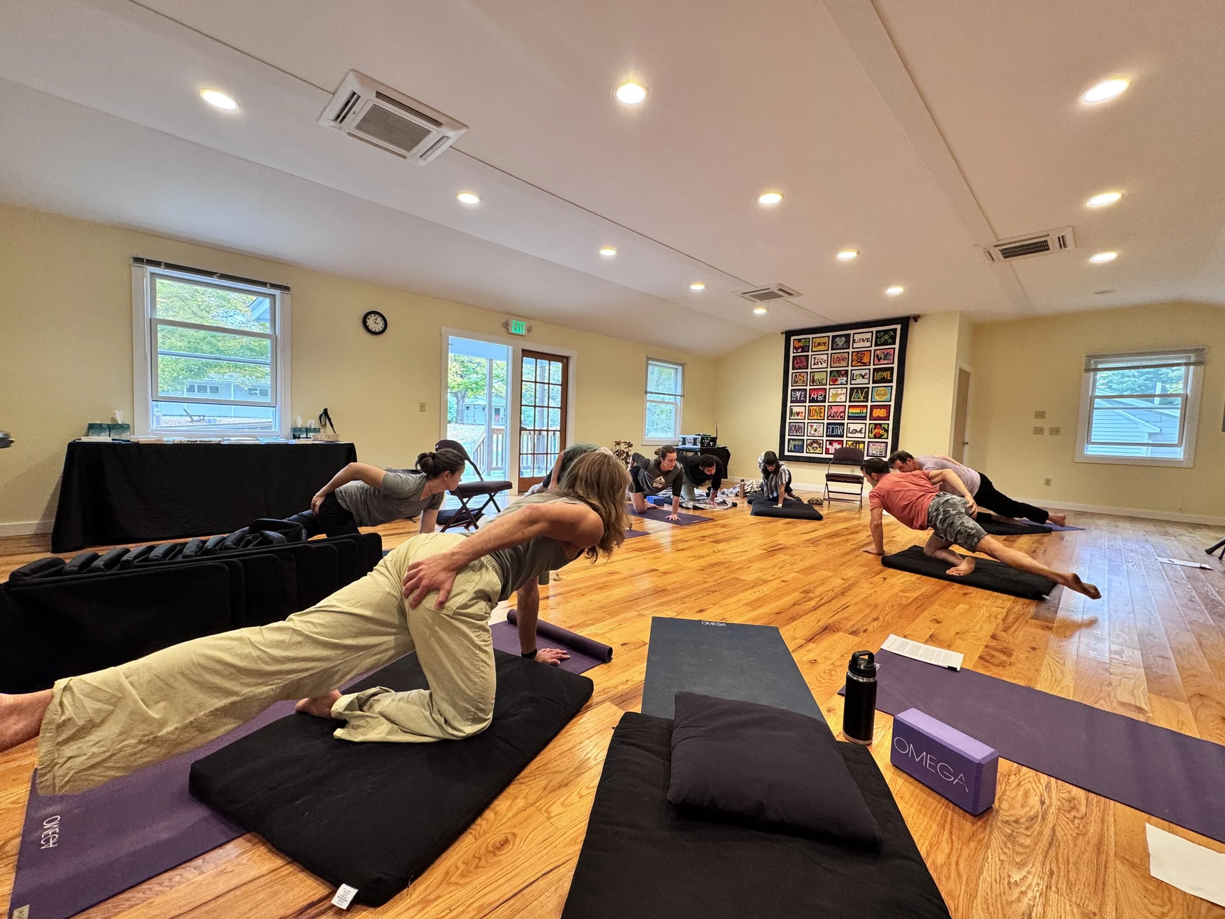 People participating in a yoga or fitness class in a spacious, well-lit room with wooden floors, windows, and colorful artwork on the wall.