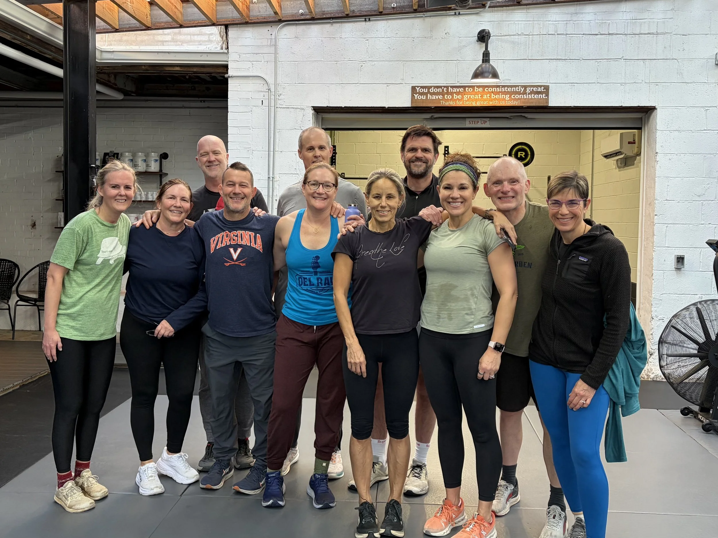 Group of thirteen people, mostly women, in workout attire, standing together in a gym, smiling for the photo.