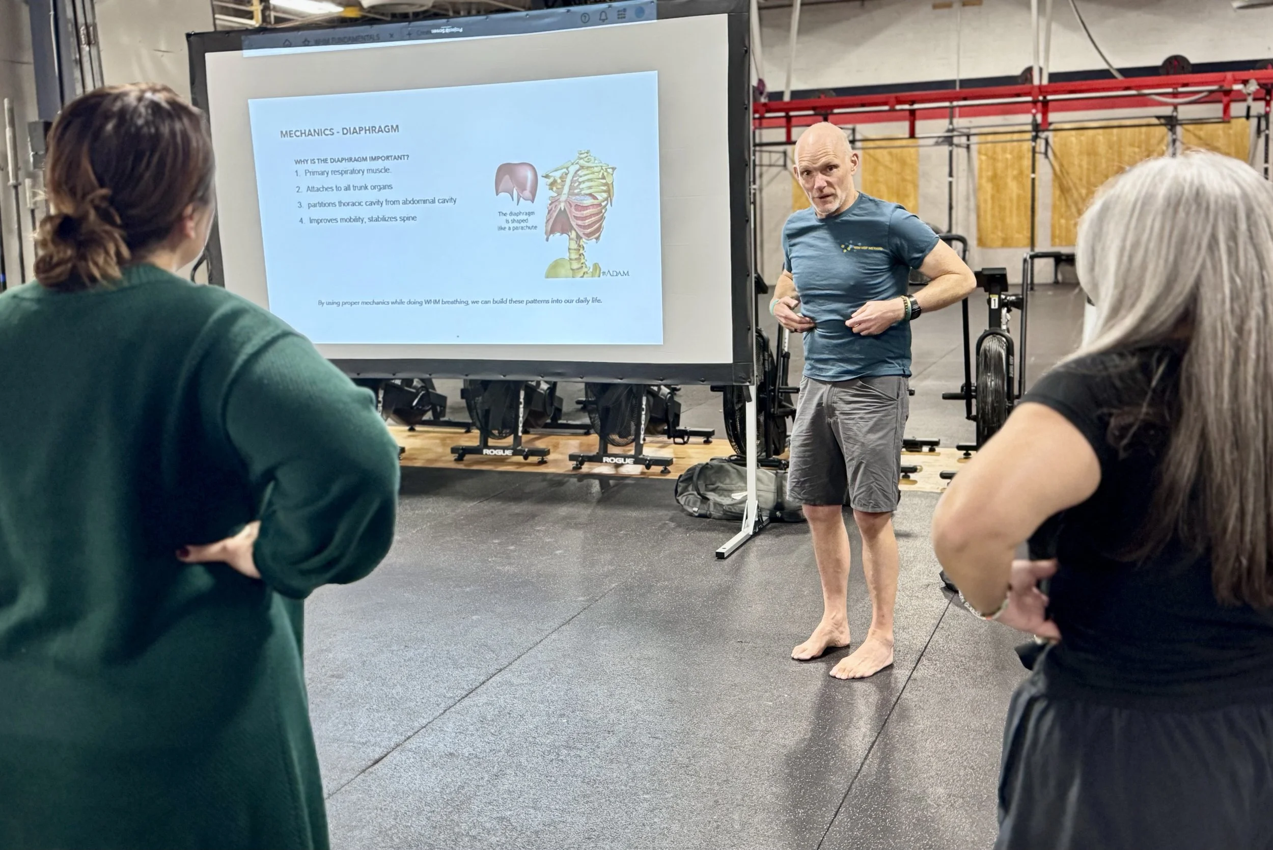 A man giving a lecture to two women in a gym, with a presentation slide about diaphragm mechanics displayed on a large screen.