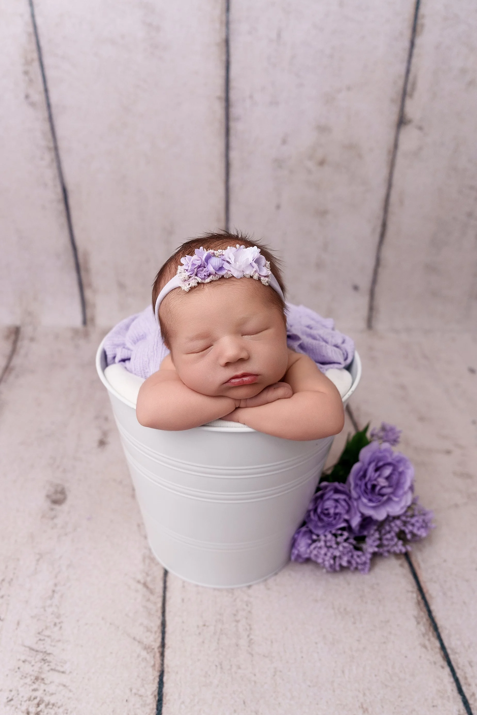 Peacefully sleeping newborn baby girl resting her chin on folded hands in a white bucket with lavender flowers, photographed at a studio serving Granite City, Illinois.