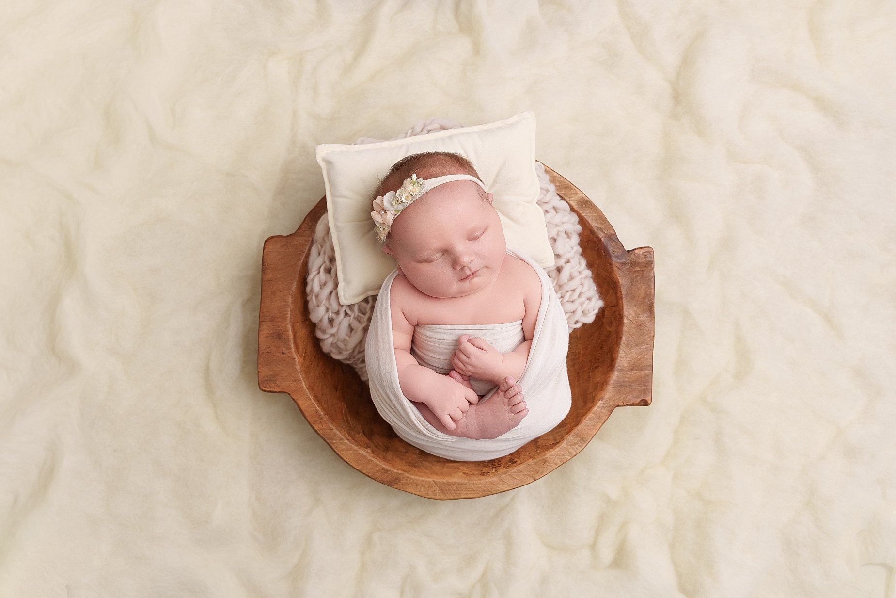 Sleeping newborn baby girl wearing an ivory wrap and delicate floral headband, posed in a wooden bowl on a soft cream backdrop at a Glen Carbon, Illinois photography studio.