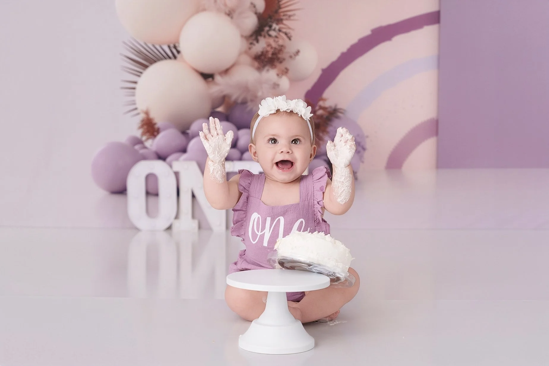 Smiling one-year-old girl with frosting-covered hands during a purple cake smash photography session near Edwardsville, Illinois.