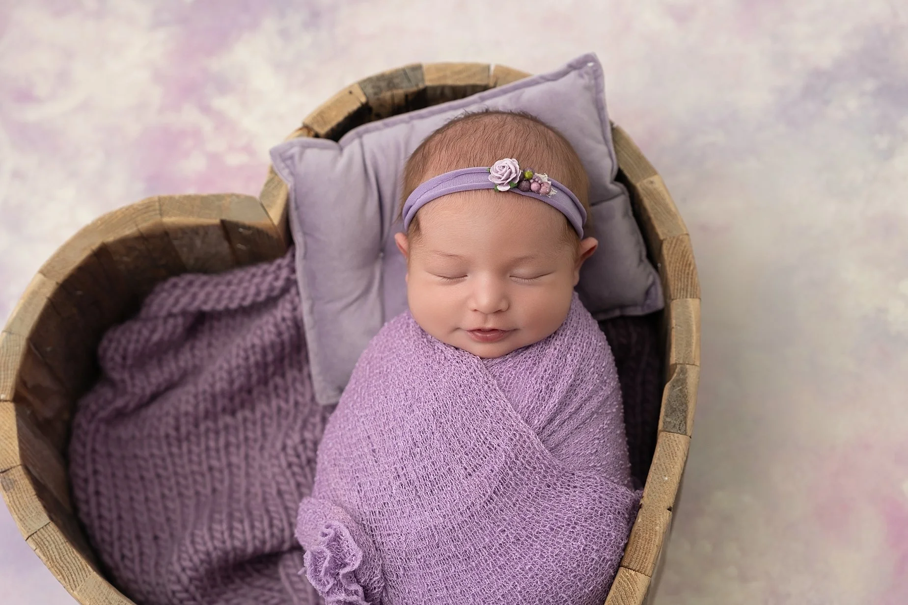 Sleeping newborn baby girl wrapped in soft lavender fabric with matching floral headband in a wooden prop during newborn photography session at Conner James Photography in Glen Carbon IL.
