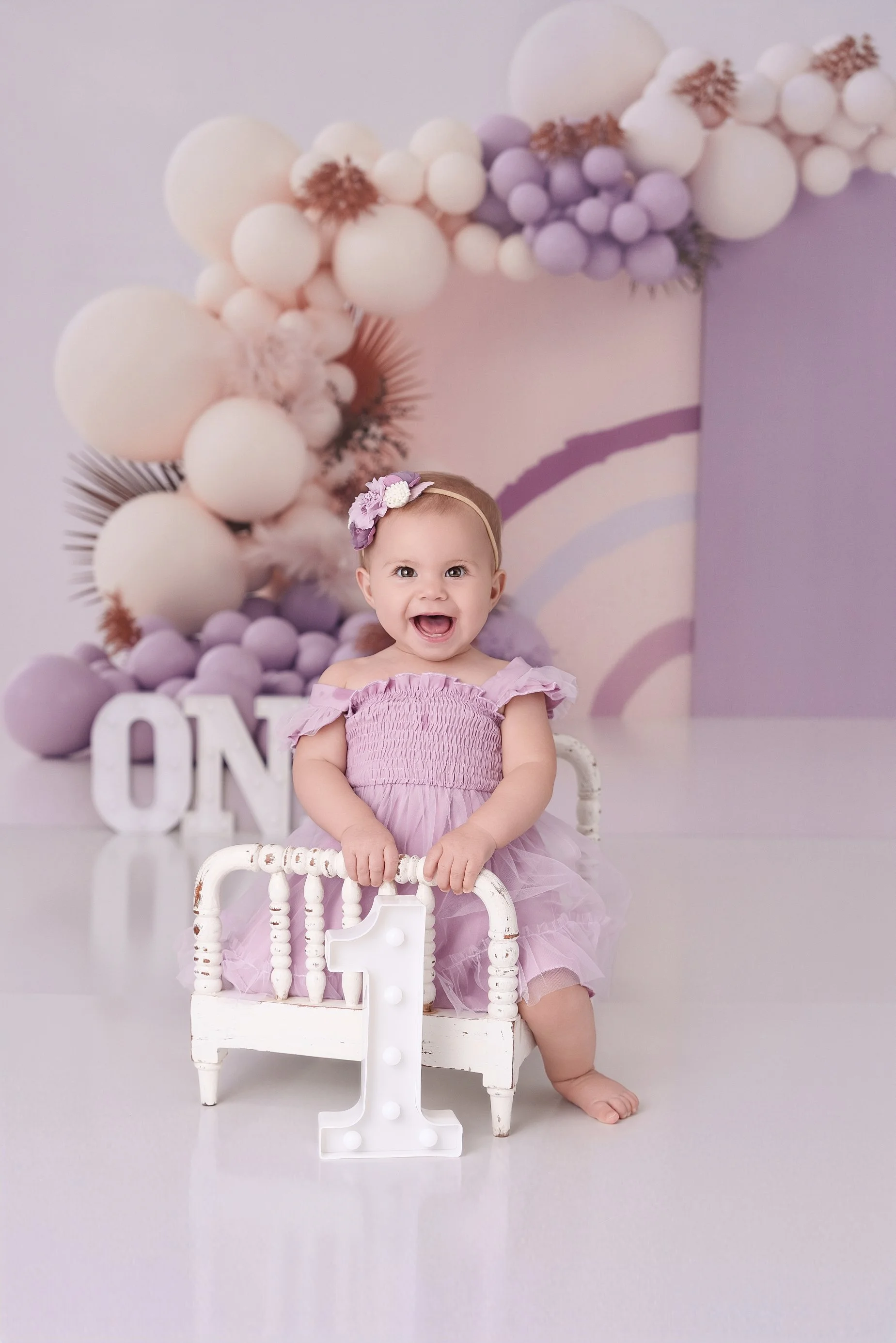 Smiling one-year-old girl in a lavender dress posing with a number one during a first birthday photography session near Edwardsville, IL.
