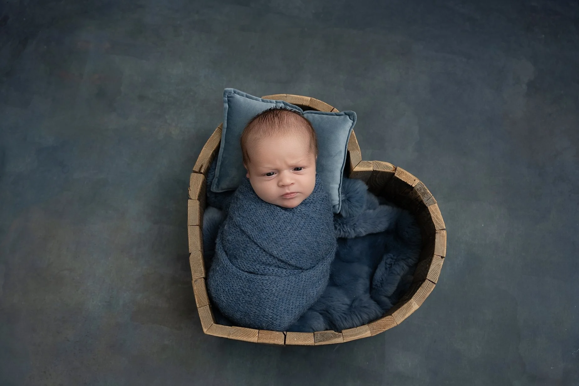 Newborn baby boy wrapped in blue fabric posing in a wooden bucket during a studio newborn photography session by an Edwardsville IL newborn photographer.