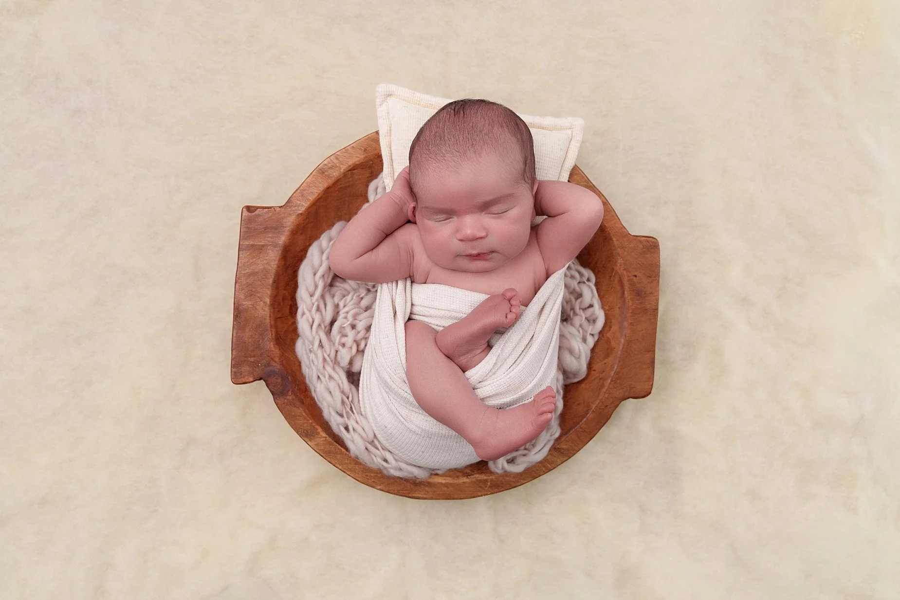 Overhead newborn portrait of a sleeping baby resting in a rustic wooden bowl with cream wraps and soft knitted textures at a Glen Carbon, IL photography studio.