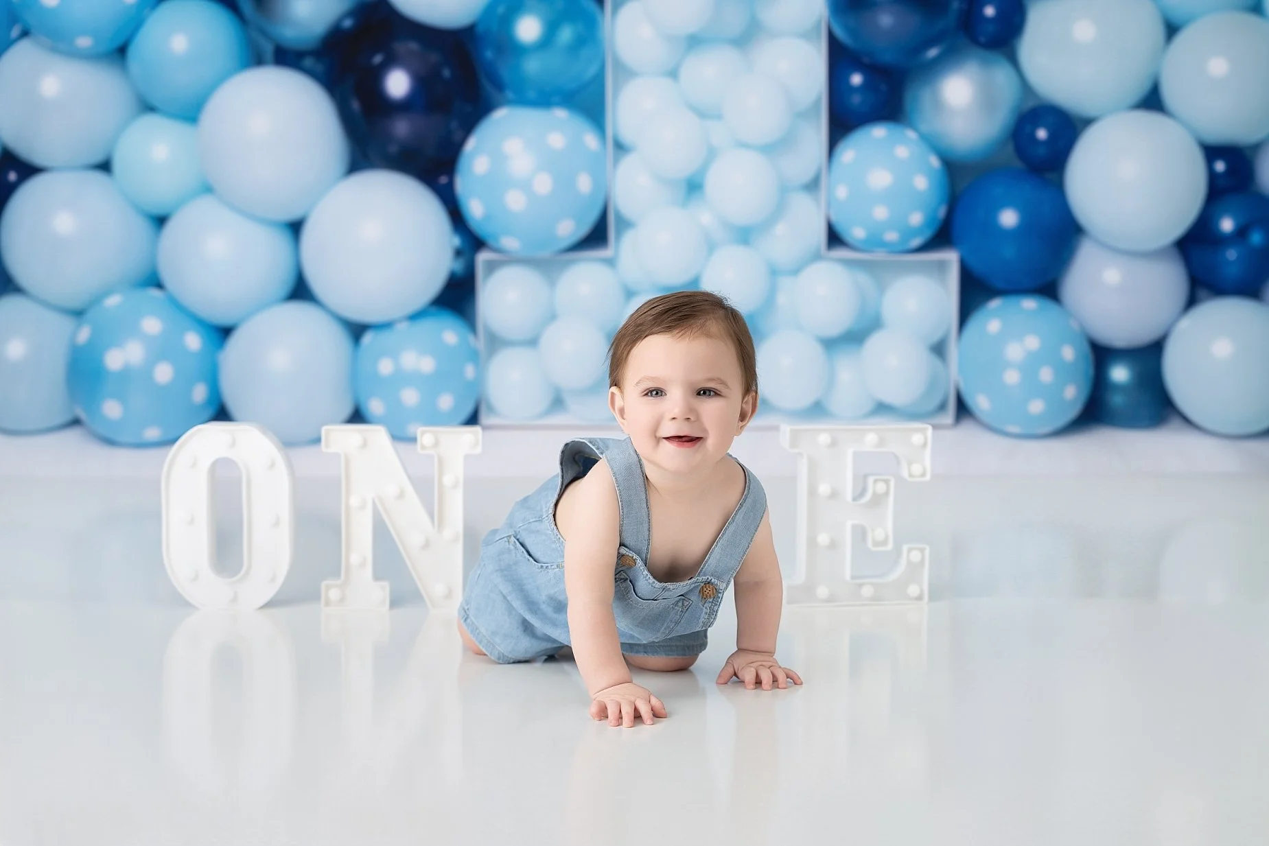 One year old baby boy crawling during milestone photography session with blue first birthday balloon setup.