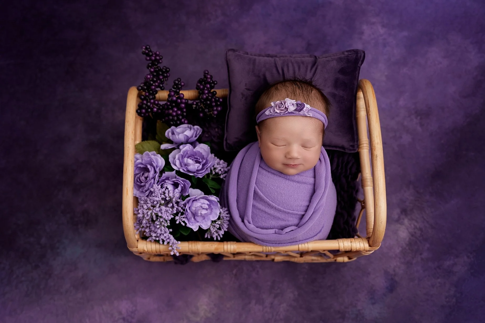 Sleeping newborn baby girl wrapped in lavender inside a wooden cradle with purple flowers, photographed by a newborn photographer serving Edwardsville, IL.