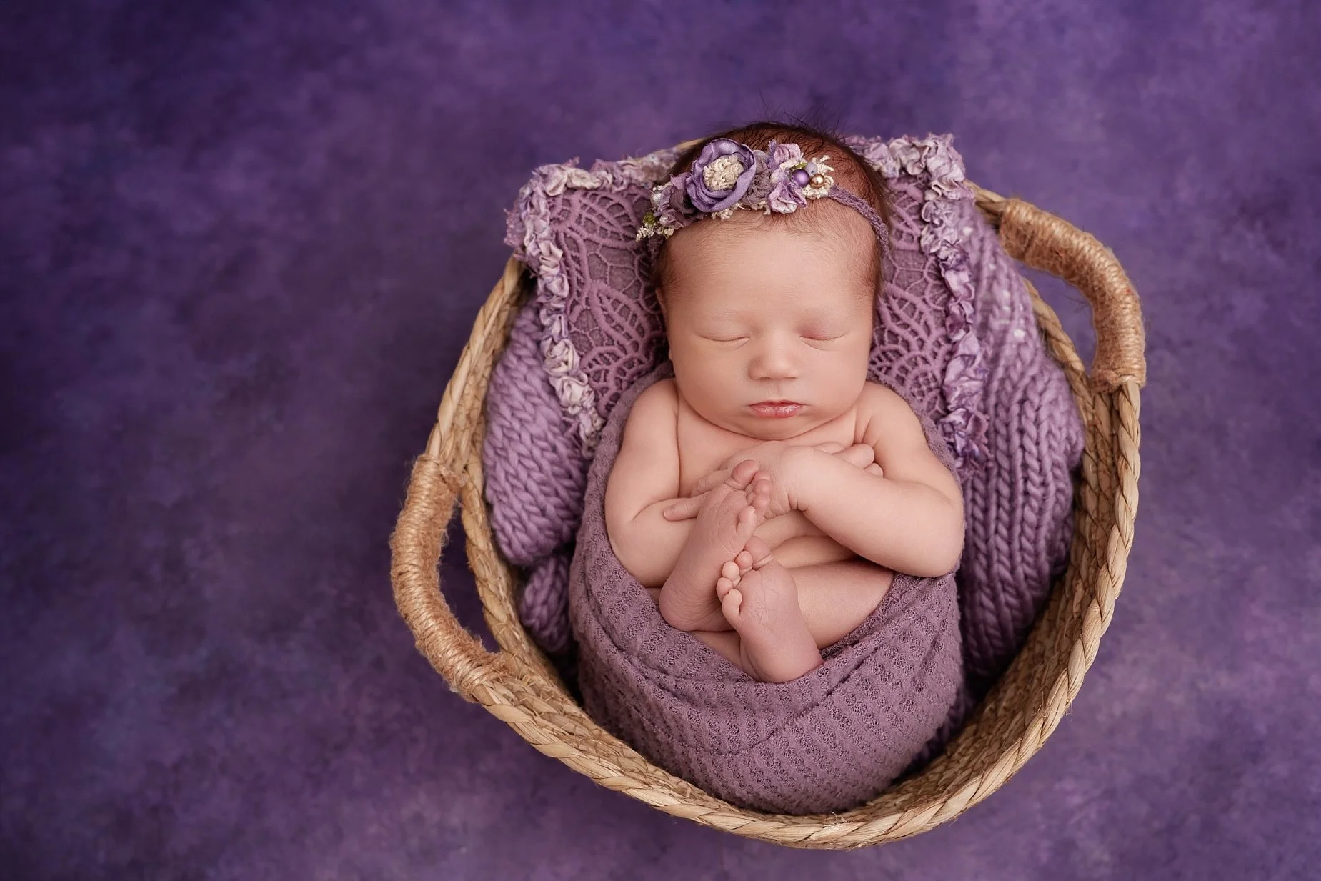 Sleeping newborn baby girl wearing a purple floral headband, posed with her tiny feet together in a woven basket with lavender blankets at a Glen Carbon, IL photography studio.