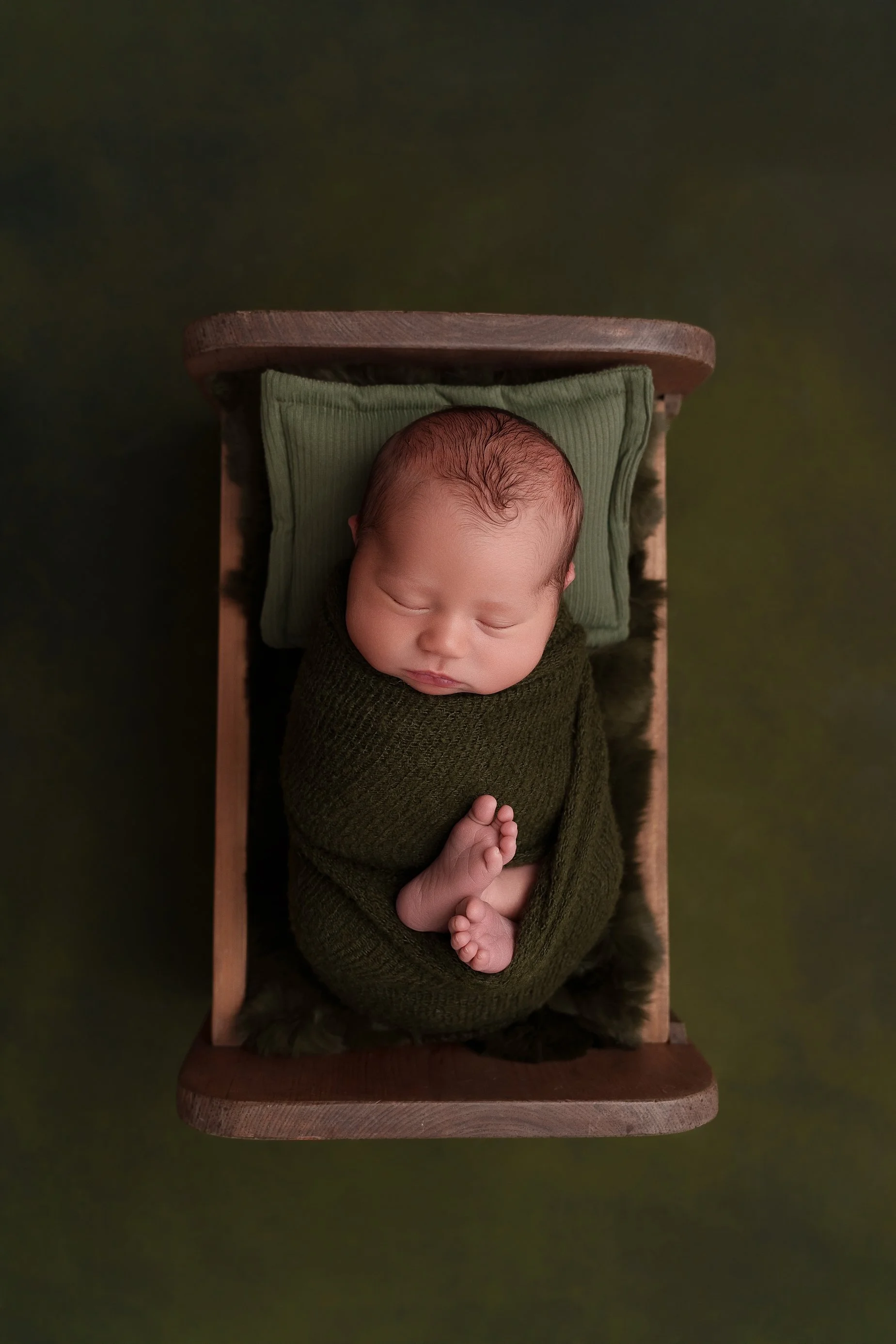 Newborn baby wrapped in olive green in a wooden bed during a newborn photography session at Conner James Photography in Glen Carbon, IL.