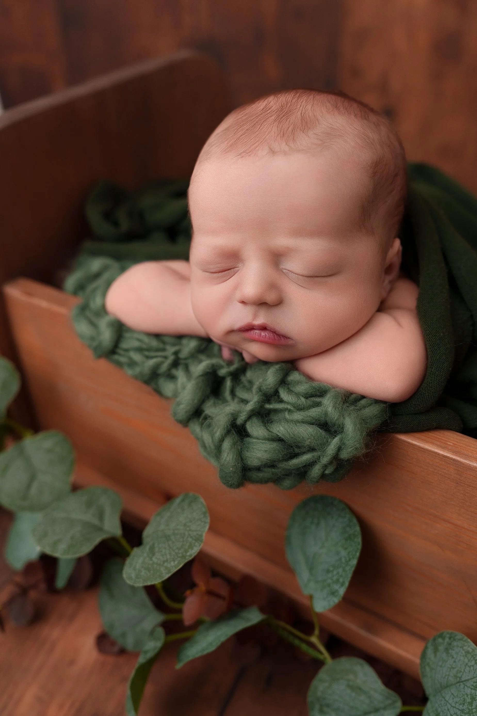 Sleeping newborn baby posed with deep green textures and a wooden prop during an Edwardsville area newborn photography session.