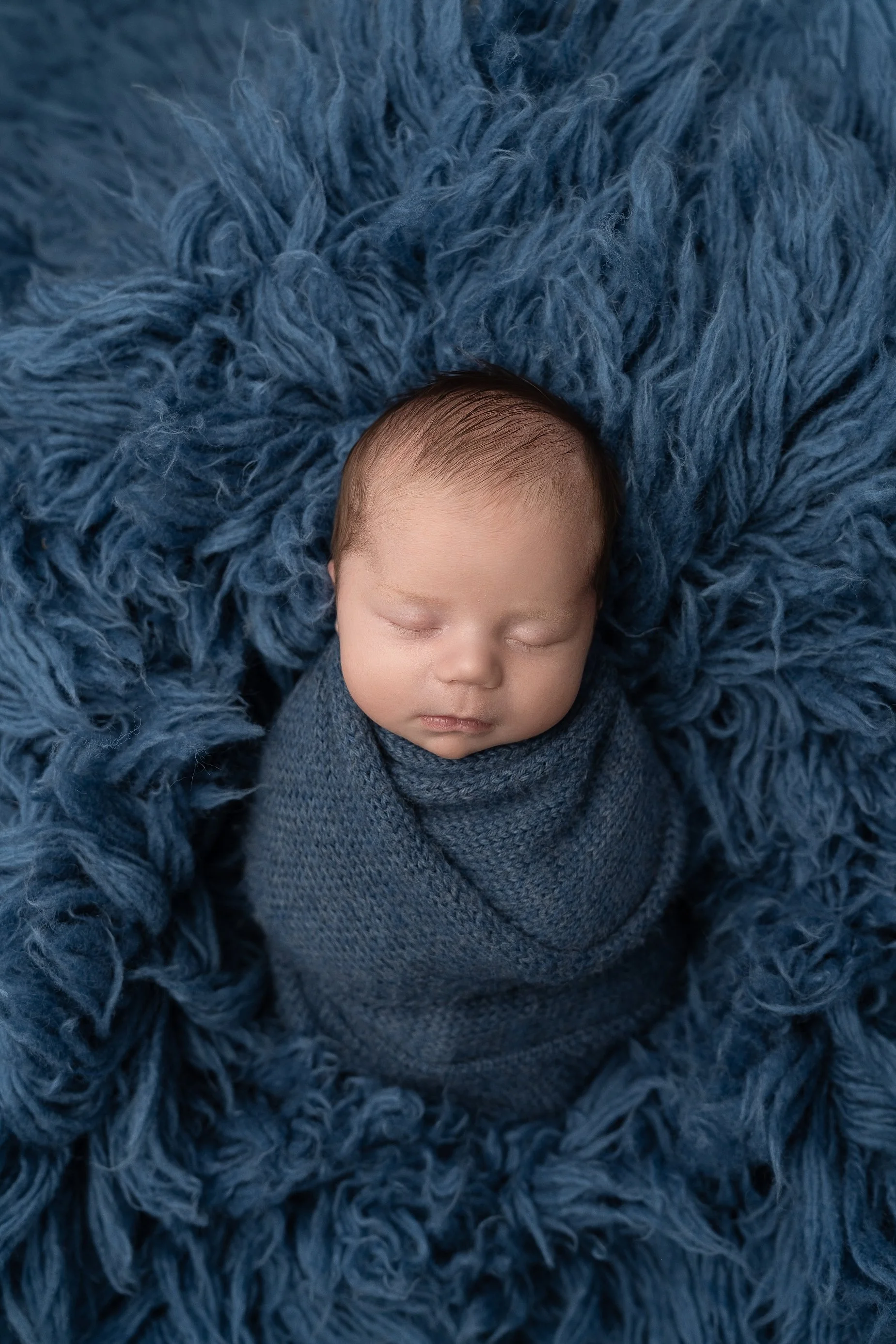 Sleeping newborn baby boy wrapped in blue blanket on textured backdrop Glen Carbon IL.