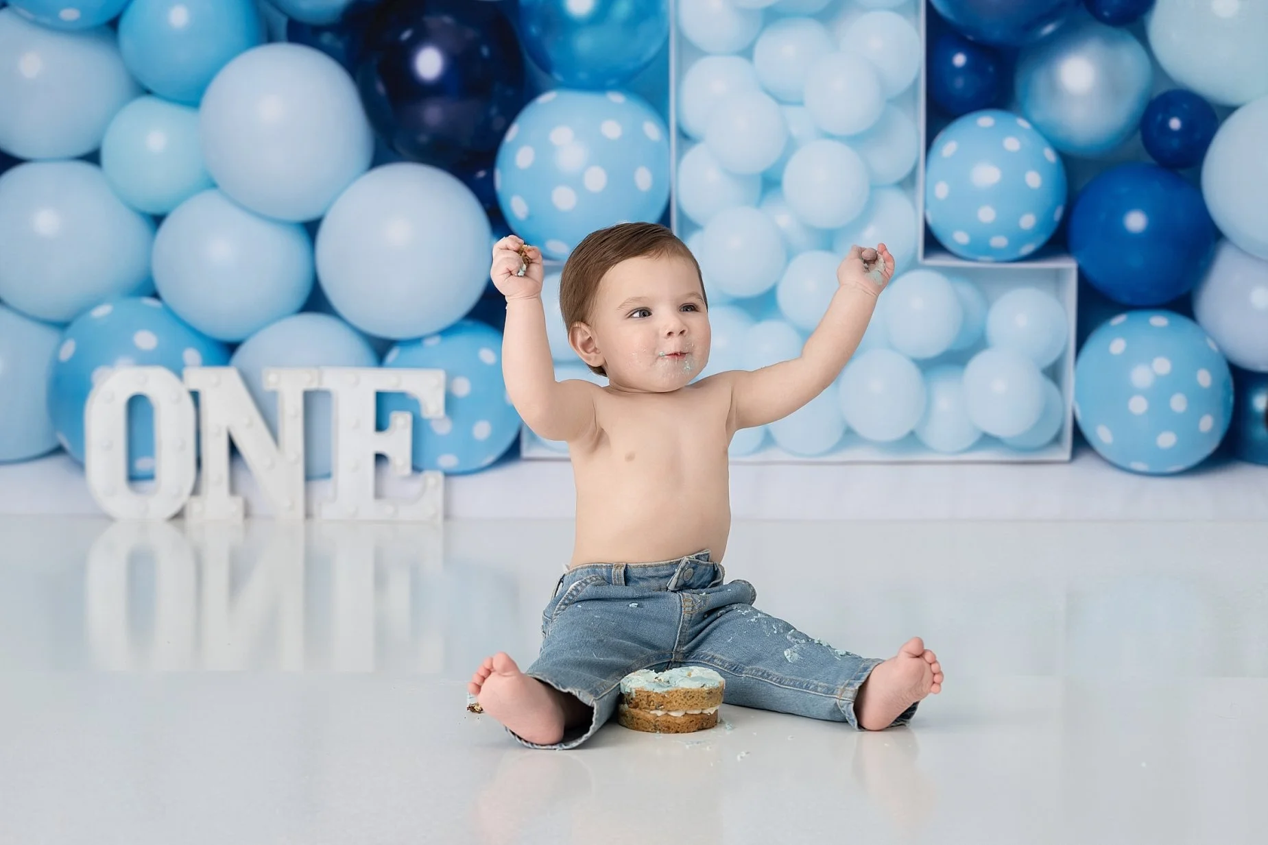 One year old baby boy celebrating his first birthday cake smash with blue balloon decorations and birthday cake.