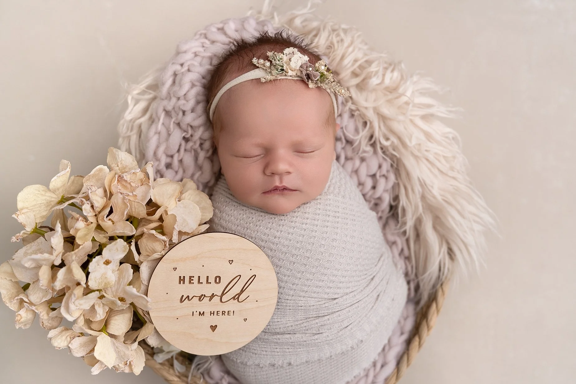 Sleeping newborn baby girl wrapped in cream fabric inside a basket with neutral florals and a Hello World wooden announcement disc.