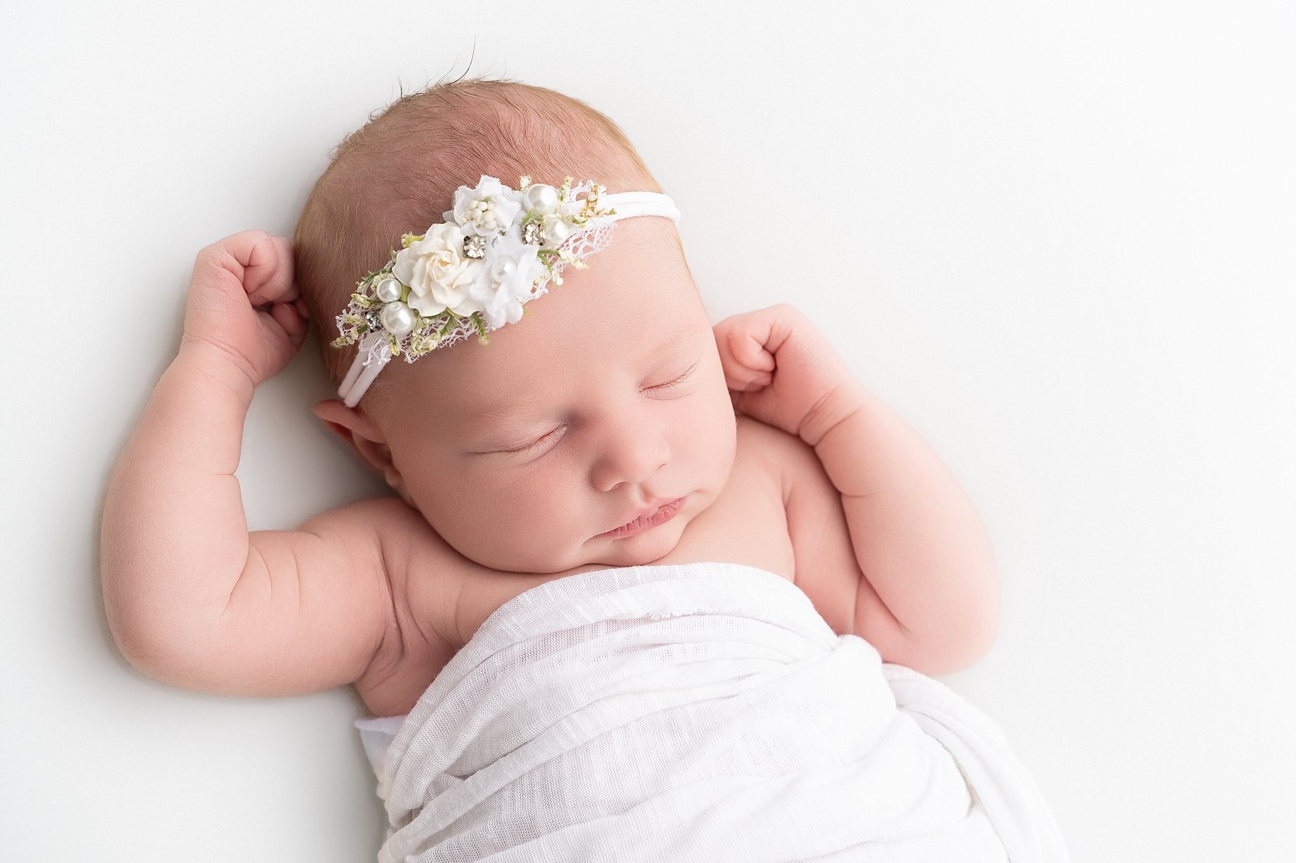 Close-up portrait of newborn baby Emilia sleeping on a white backdrop wearing a floral headband.