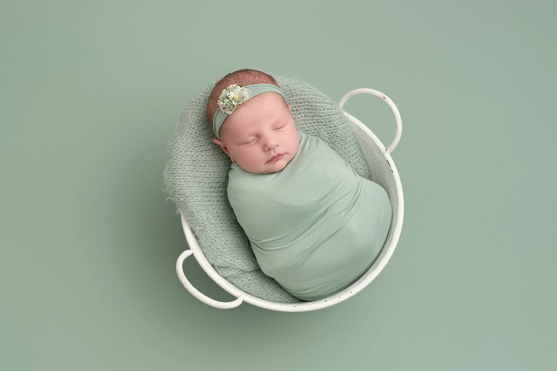 Sleeping newborn baby girl Emilia wrapped in sage green fabric in a white bowl during a professional newborn photography session.