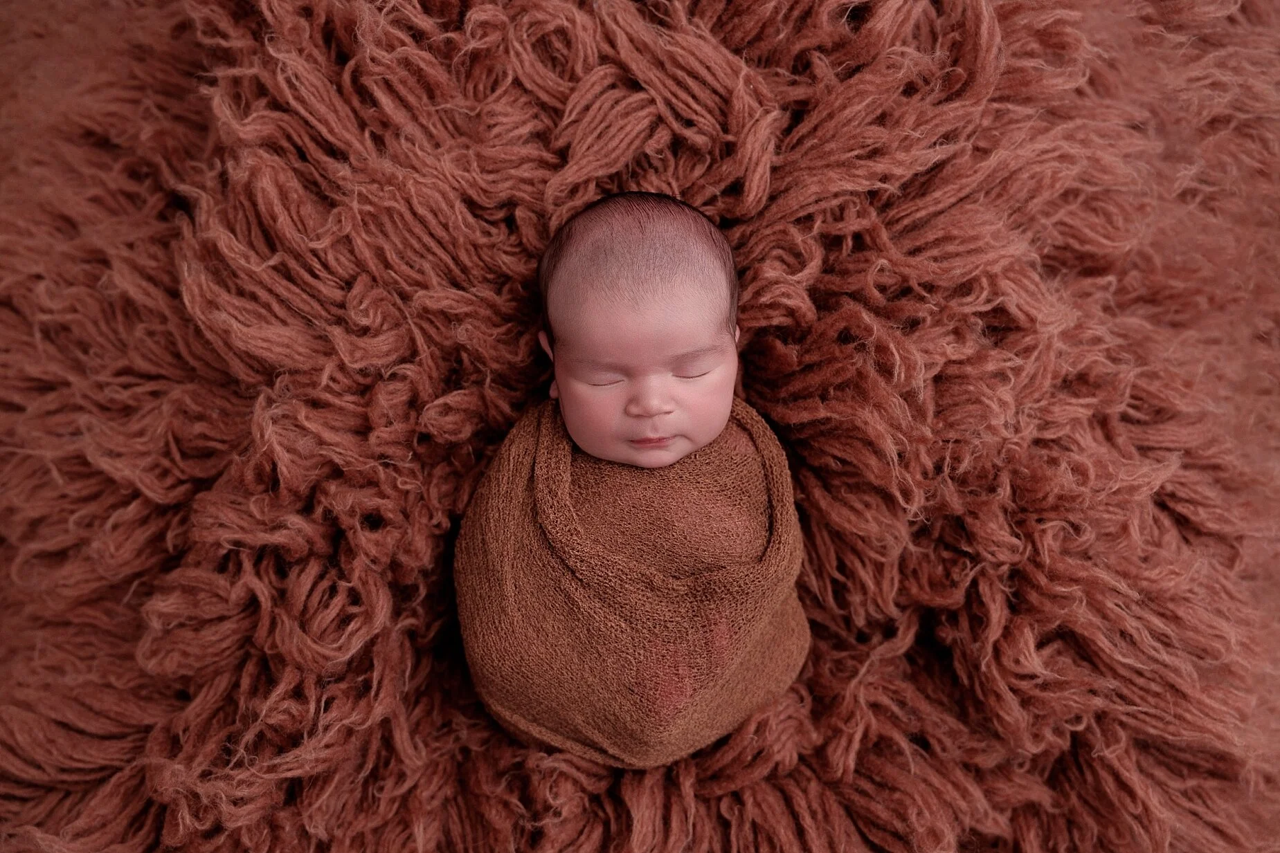 Sleeping newborn baby wrapped in a terracotta-colored blanket on a textured rust backdrop, photographed by Conner James Photography near Edwardsville, IL.
