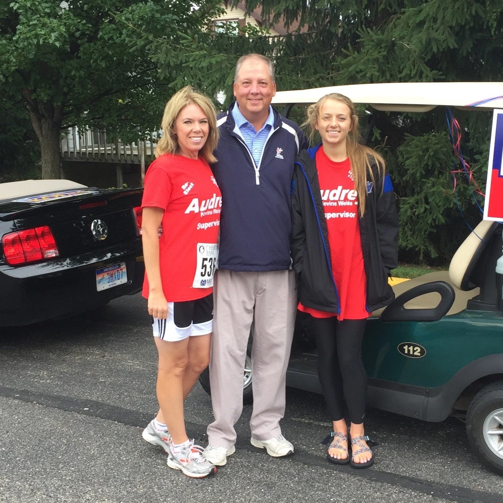 Three people standing together outdoors near a golf cart, smiling at the camera, with trees and a house in the background.