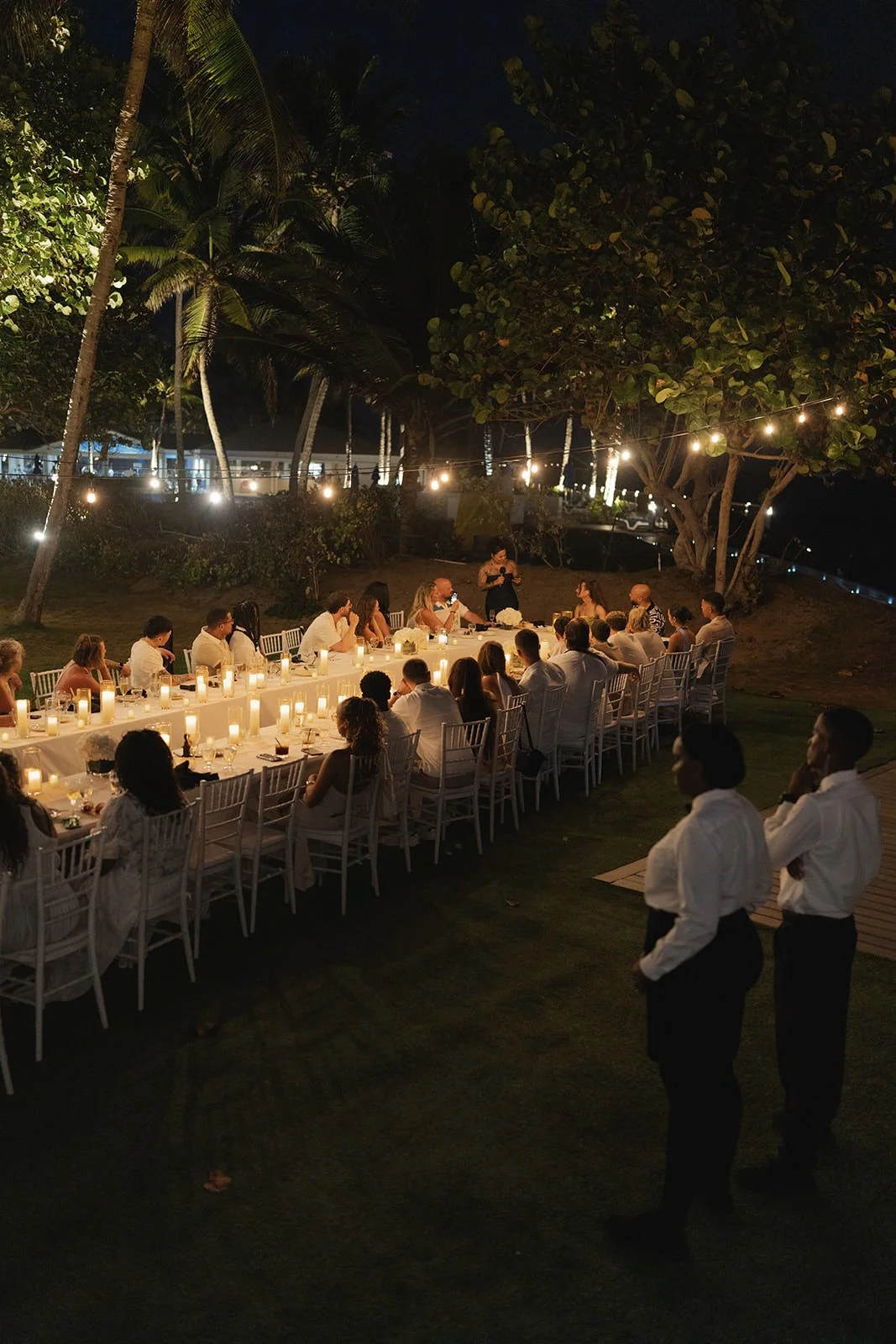 Long communal dinner table set under palm trees and string lights at night, St. Lucia wedding weekend
