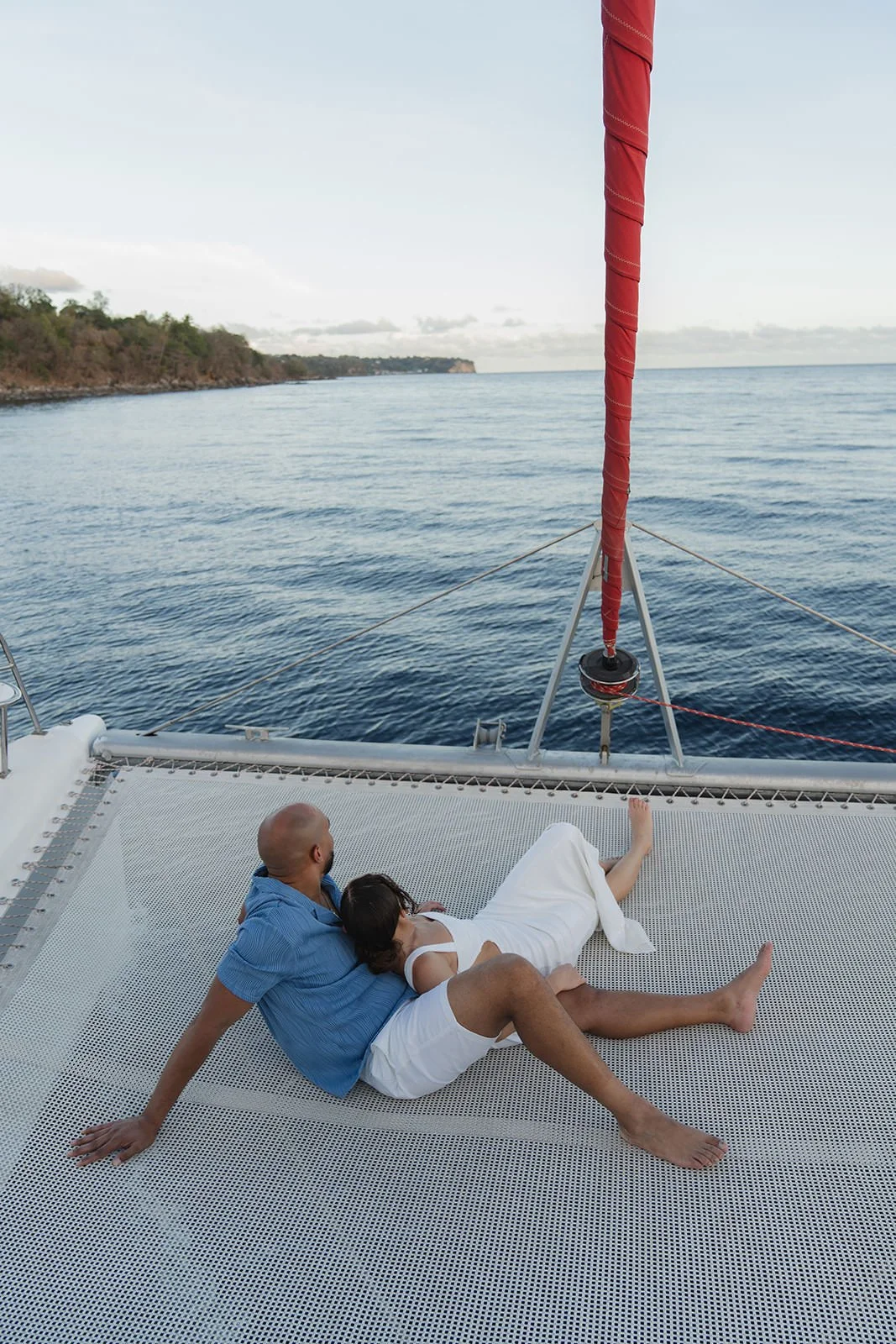 Groom and bride lying together on the catamaran's trampoline netting, embracing while looking out at St. Lucia's coastline with a red sail mast overhead, wedding weekend sailing excursion