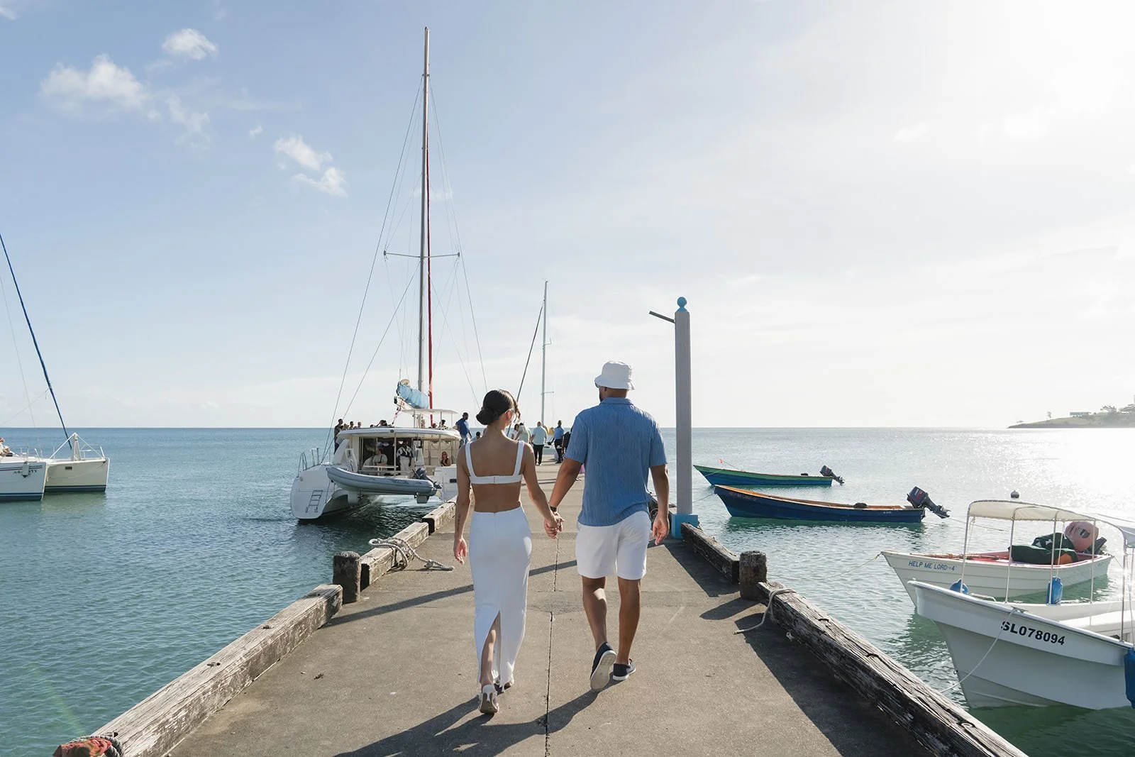 Bride in white and groom in blue walking hand in hand along a dock lined with sailboats in a St. Lucia harbor during the wedding weekend