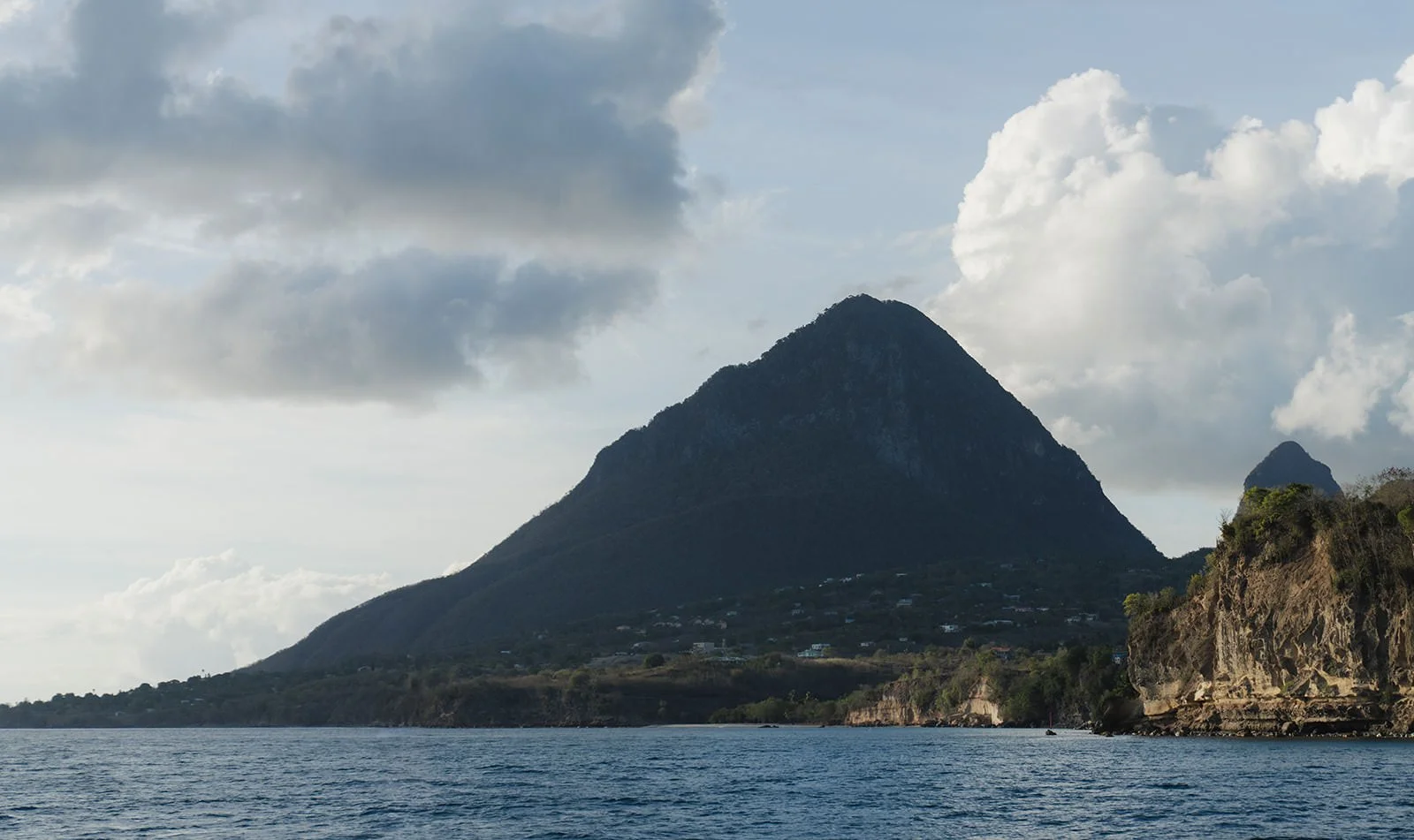 View of a St. Lucia Piton mountain peak and rocky coastline under a cloudy sky, seen from the water during the catamaran excursion