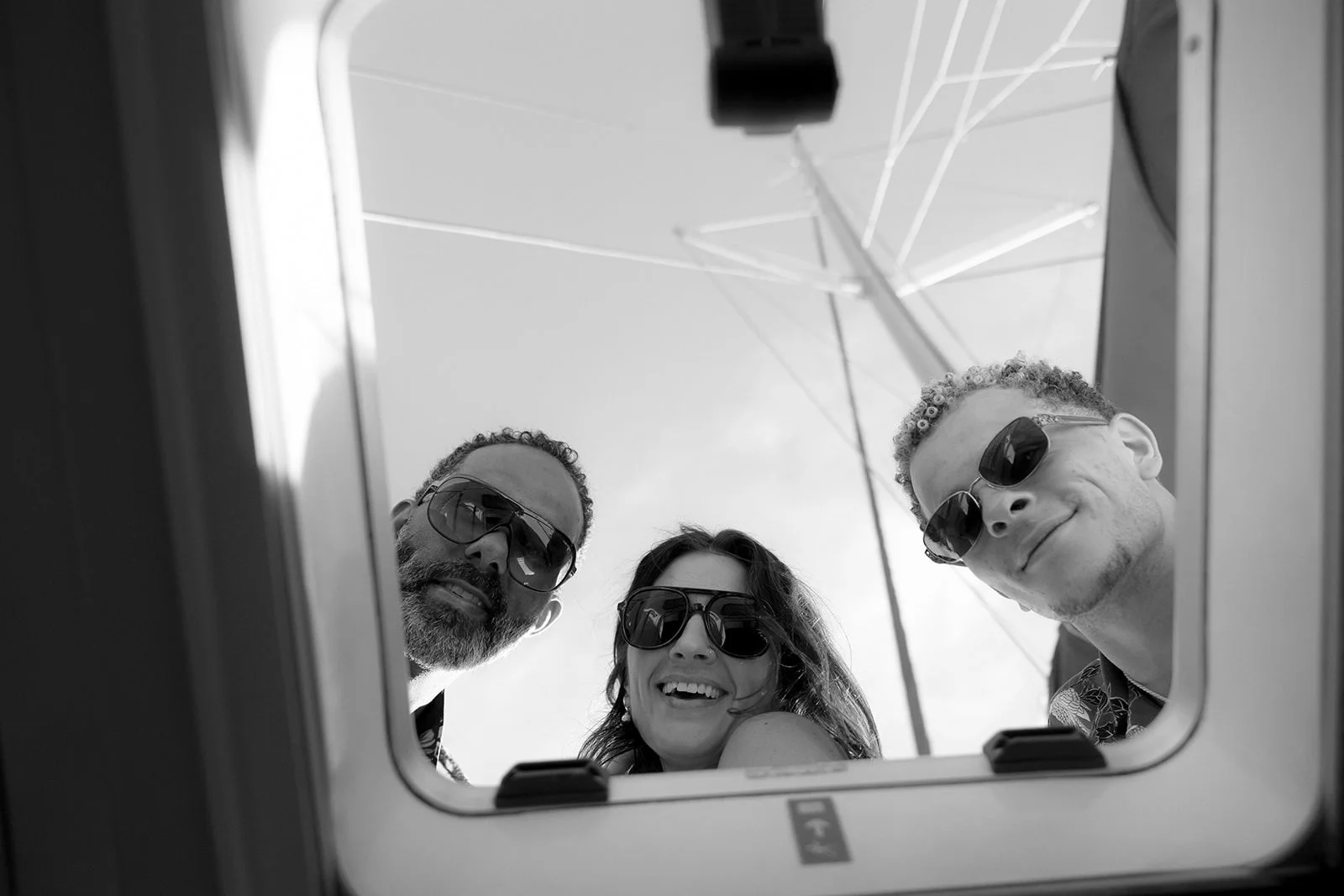 Black-and-white photo of three laughing wedding guests seen through the catamaran's window during the sailing excursion