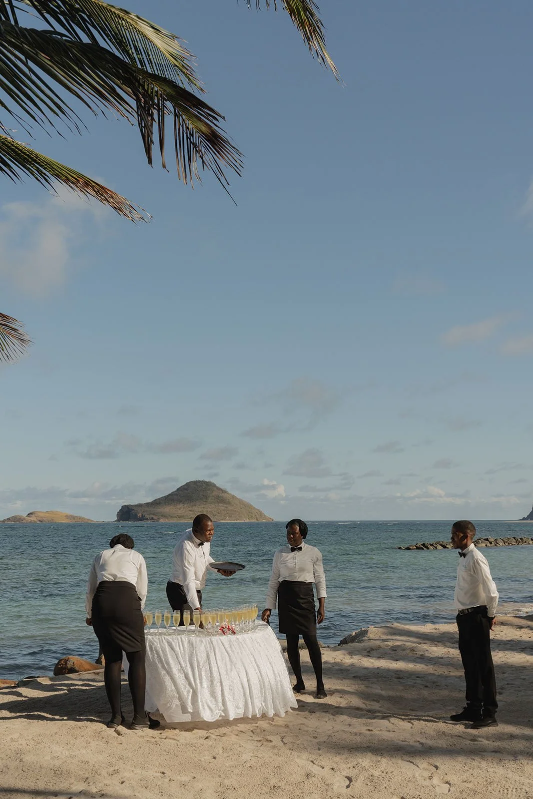 Beachside table setup with staff preparing for the wedding ceremony, a small island visible offshore in St. Lucia