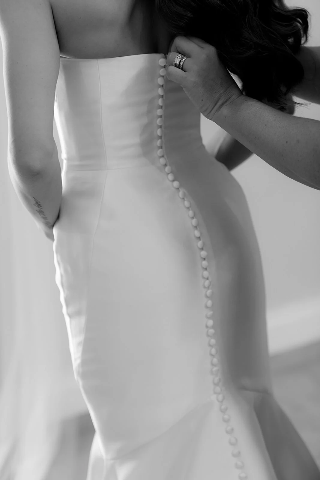 Black-and-white close-up of the back of the bride's fitted wedding gown as it is buttoned up before the ceremony